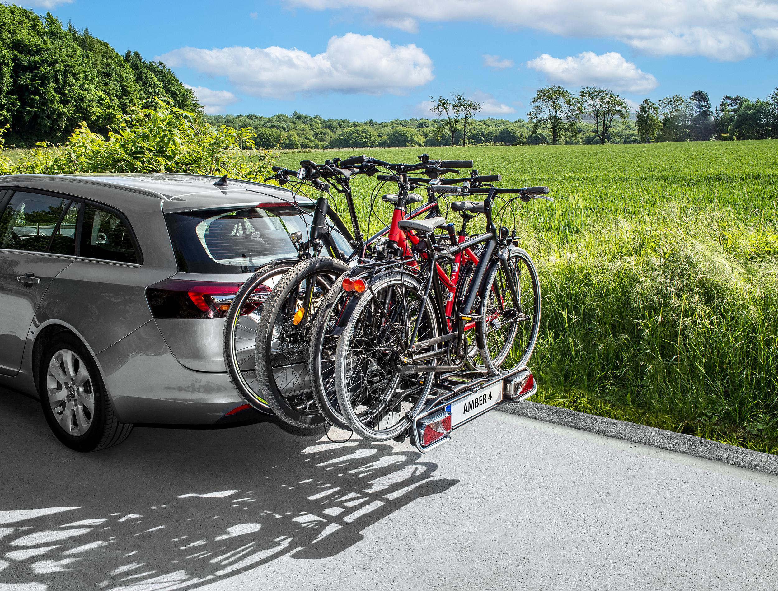 Auto mit Fahrradträger mit drei Fahrrädern auf einer Straße, grüne Felder im Hintergrund.