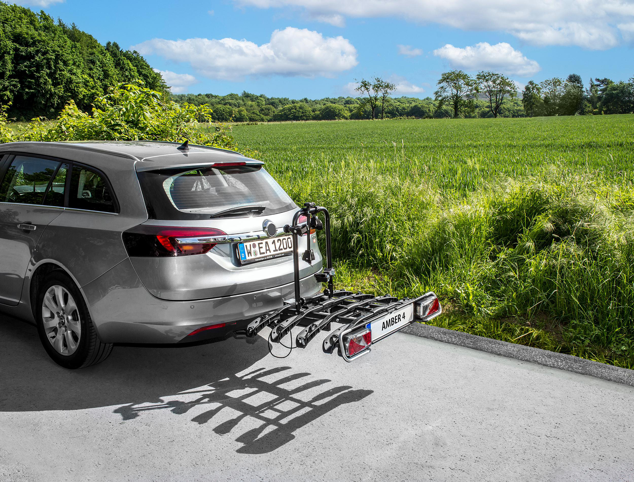 Ein silbernes Auto mit einem Fahrradträger. Das Auto parkt auf einer Straße neben einem grünen Feld und Bäumen.