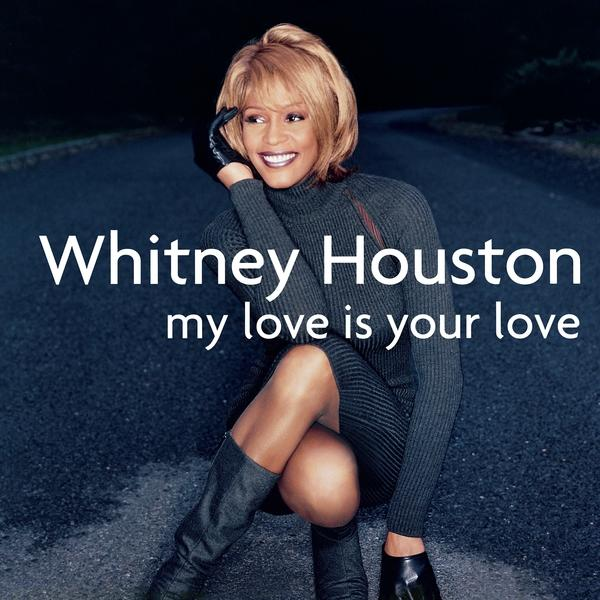 Whitney Houston egy kék-szürke úton, térdelve, fekete garbóban, mosolyogva.