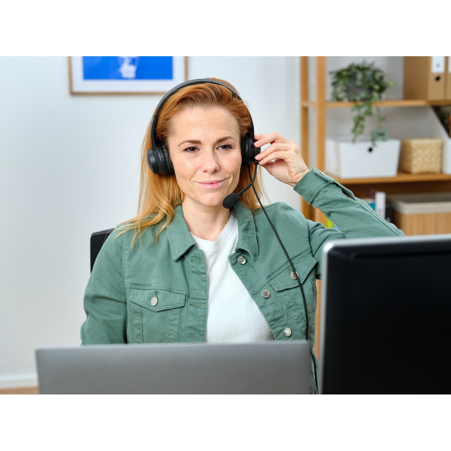 A woman wearing a headset smiles while looking at a laptop.