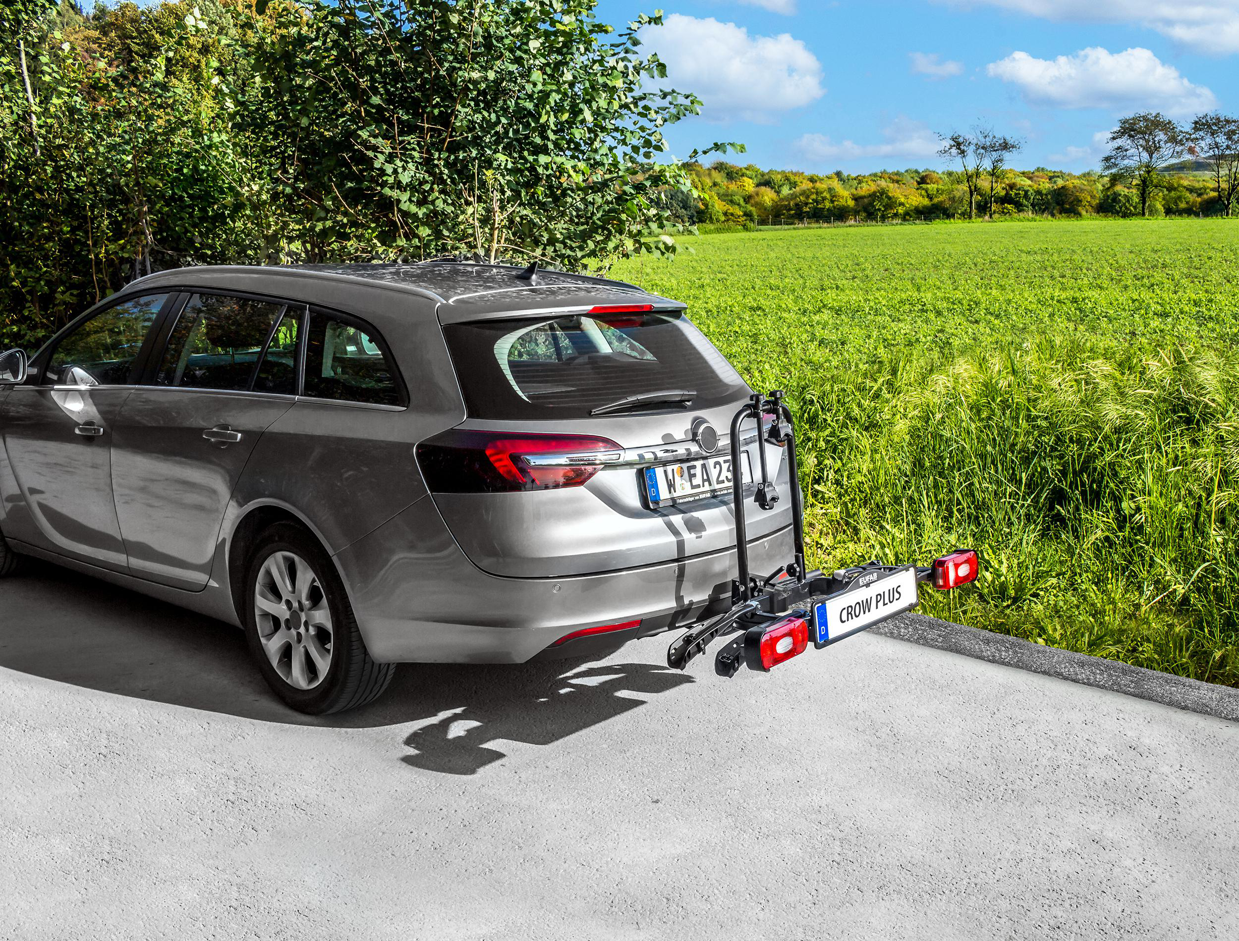 Ein graues Auto mit Fahrradträger, geparkt auf einer Straße mit grünem Feld im Hintergrund.