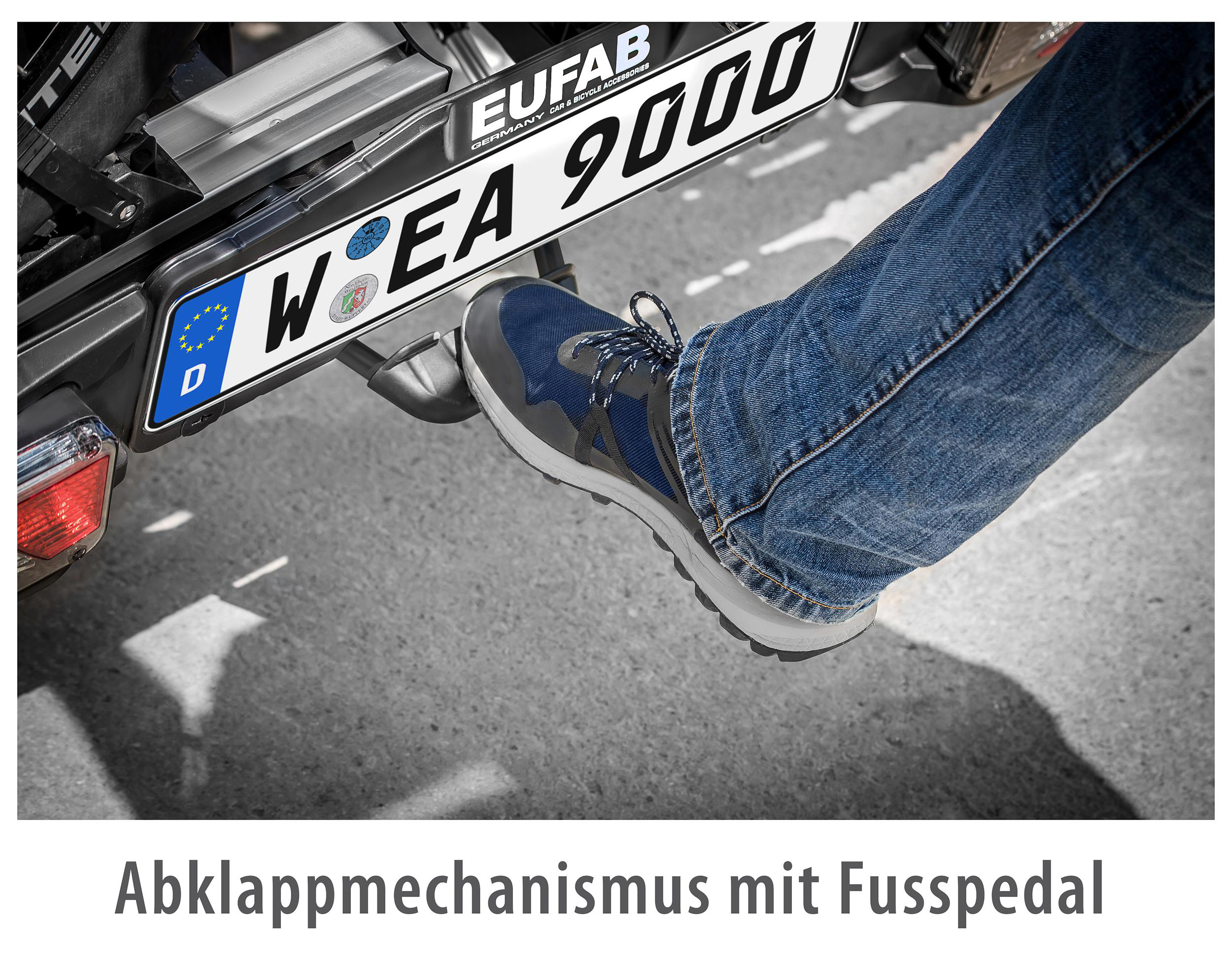 Ein Fuß tritt auf einen Mechanismus. Ein Nummernschild ist sichtbar, die Hose ist Jeans.