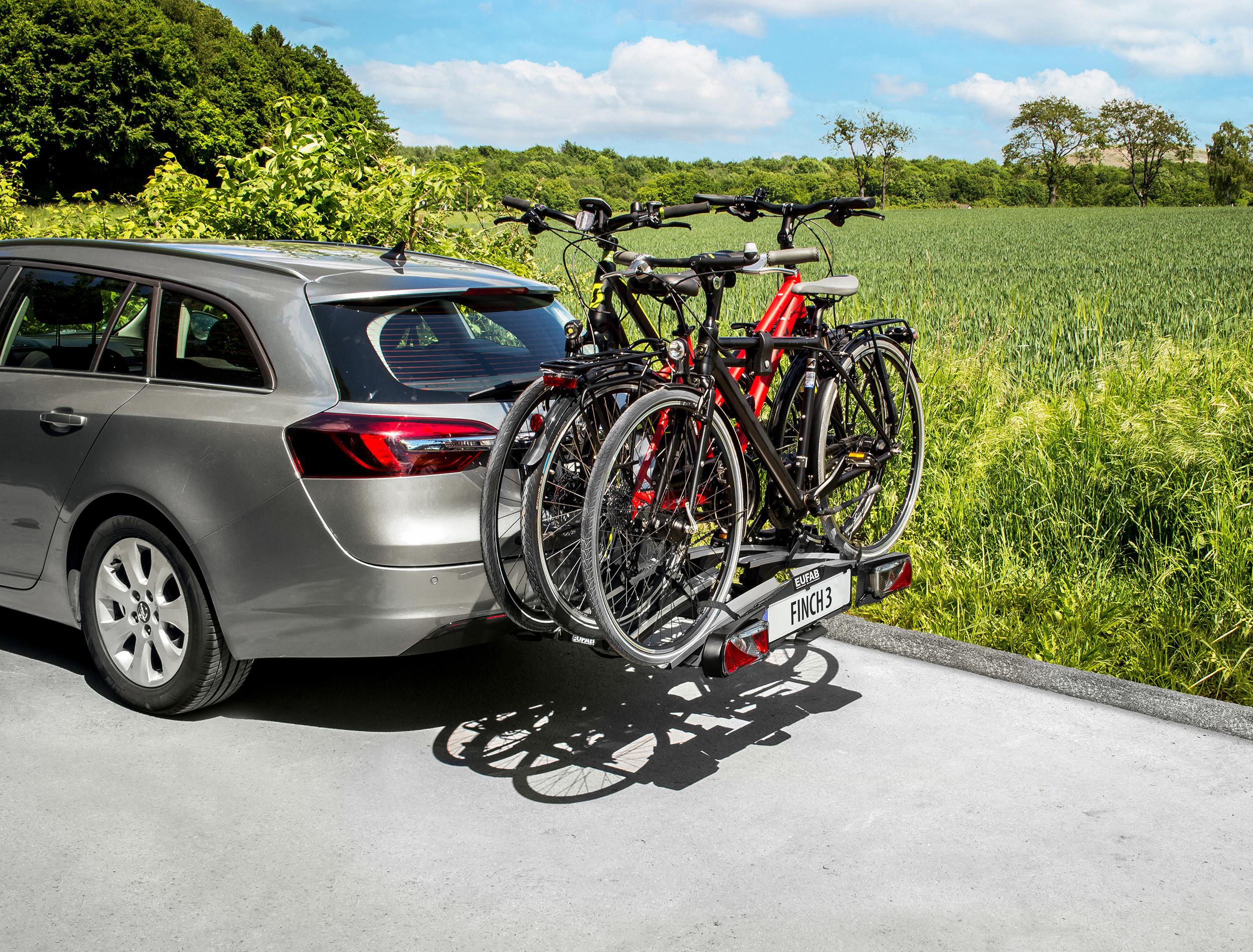 Ein graues Auto mit Fahrradträger, der drei Fahrräder trägt, geparkt auf einer Straße mit grünen Feldern.