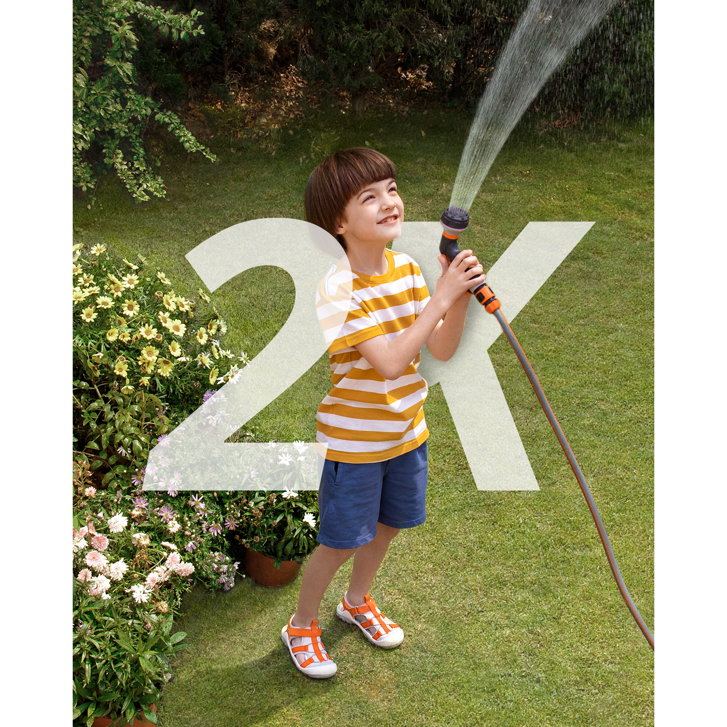 Een jongen glimlacht terwijl hij een slang vasthoudt die water sproeit in een tuin met bloemen.