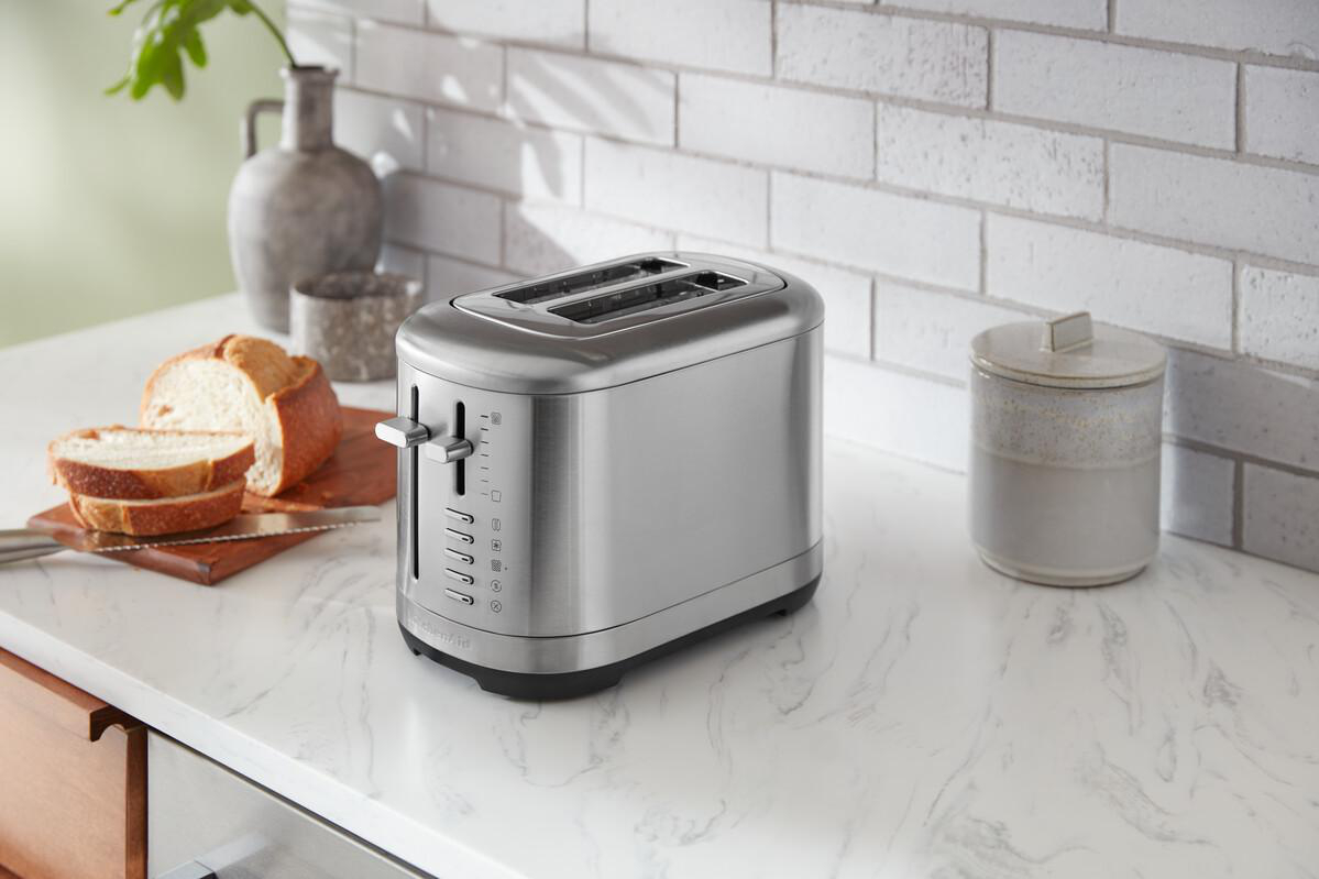 Silberner Toaster auf weißer Arbeitsplatte, mit Brot und einem Glas im Hintergrund.