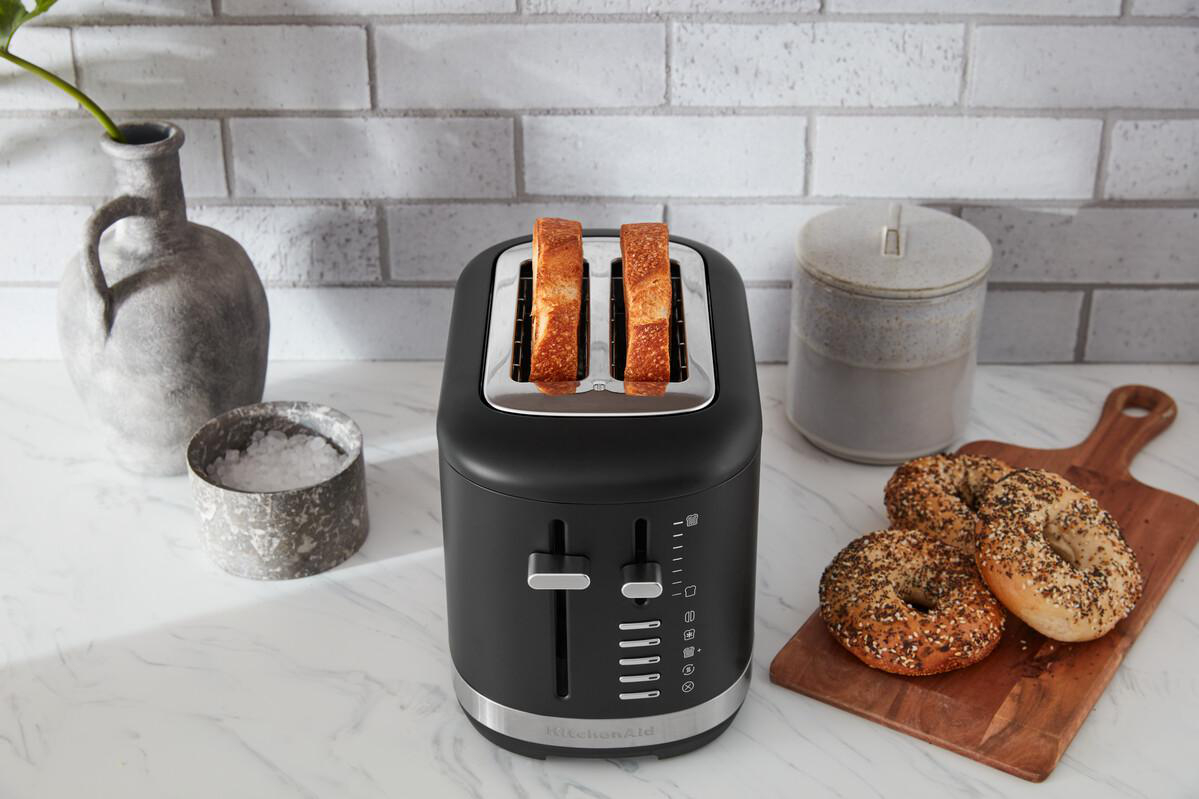 Schwarzer Toaster mit Toast in Schlitzen, Bagels auf Holzbrett und Ziegelwand im Hintergrund.