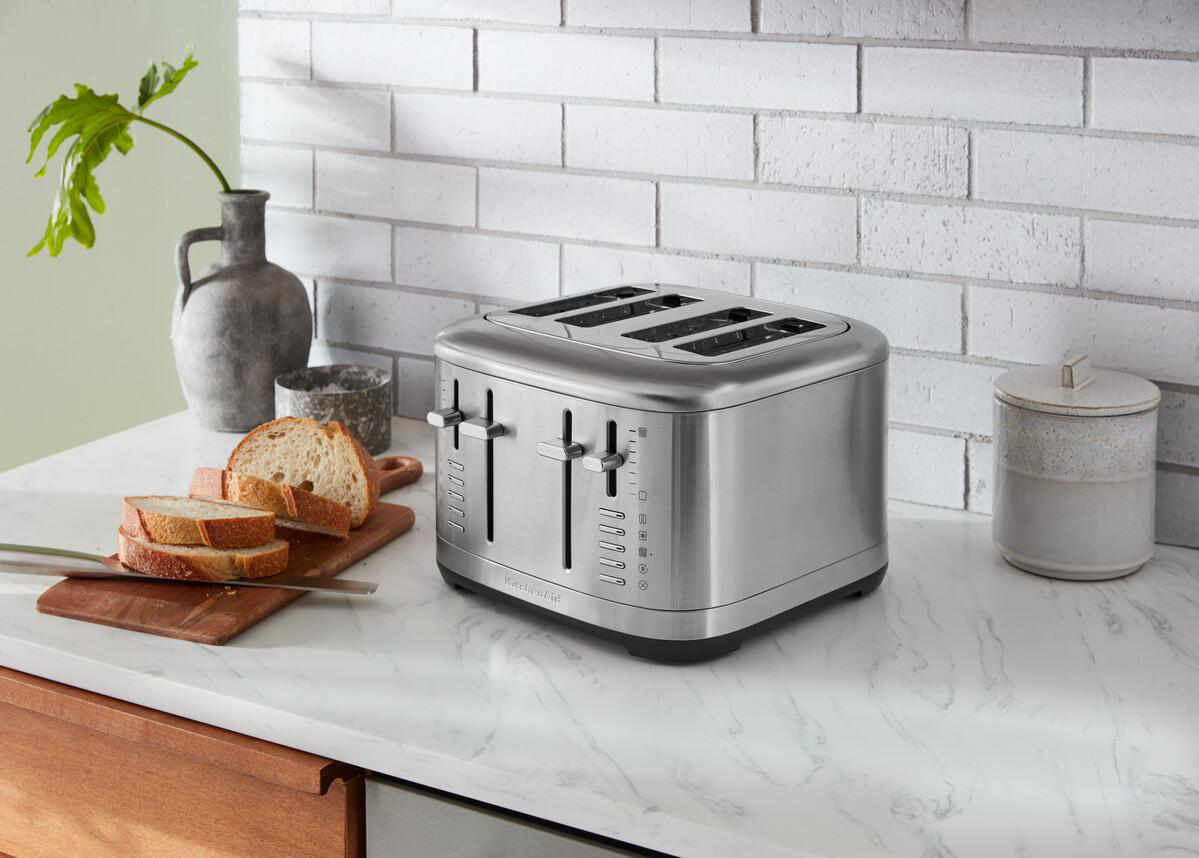 Ein silberner KitchenAid Toaster mit vier Schlitzen auf einer weißen Arbeitsplatte, mit Brot und einem Glas.