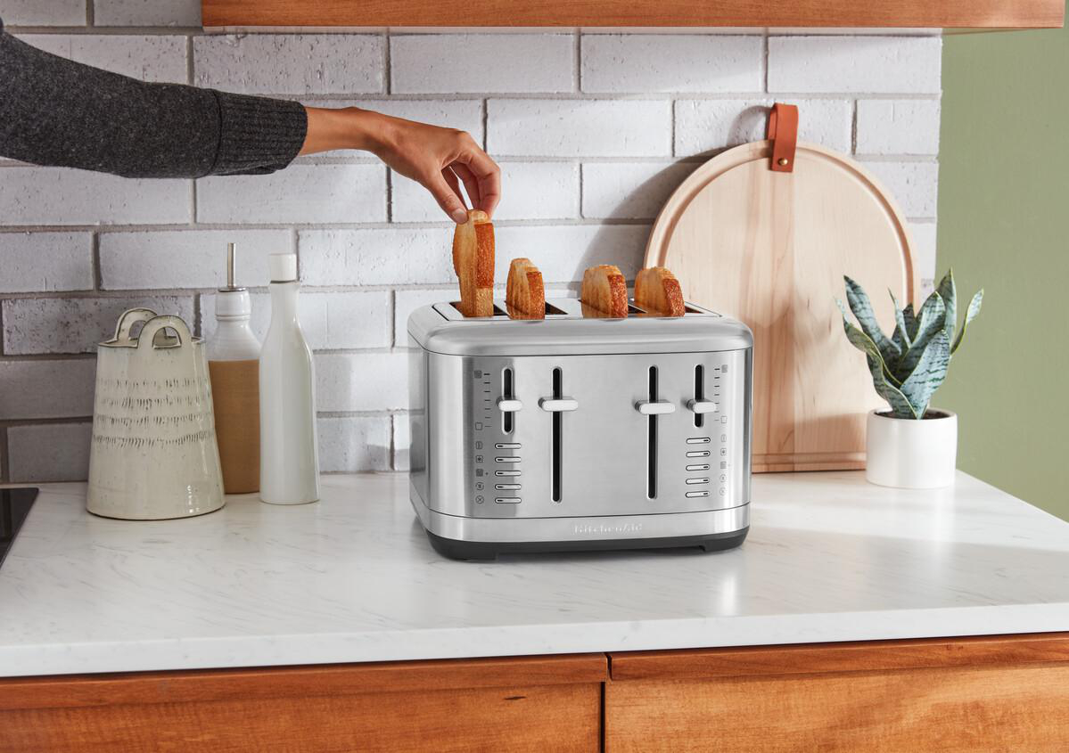 Person legt Brot in einen silbernen KitchenAid Toaster auf einer weißen Arbeitsplatte.