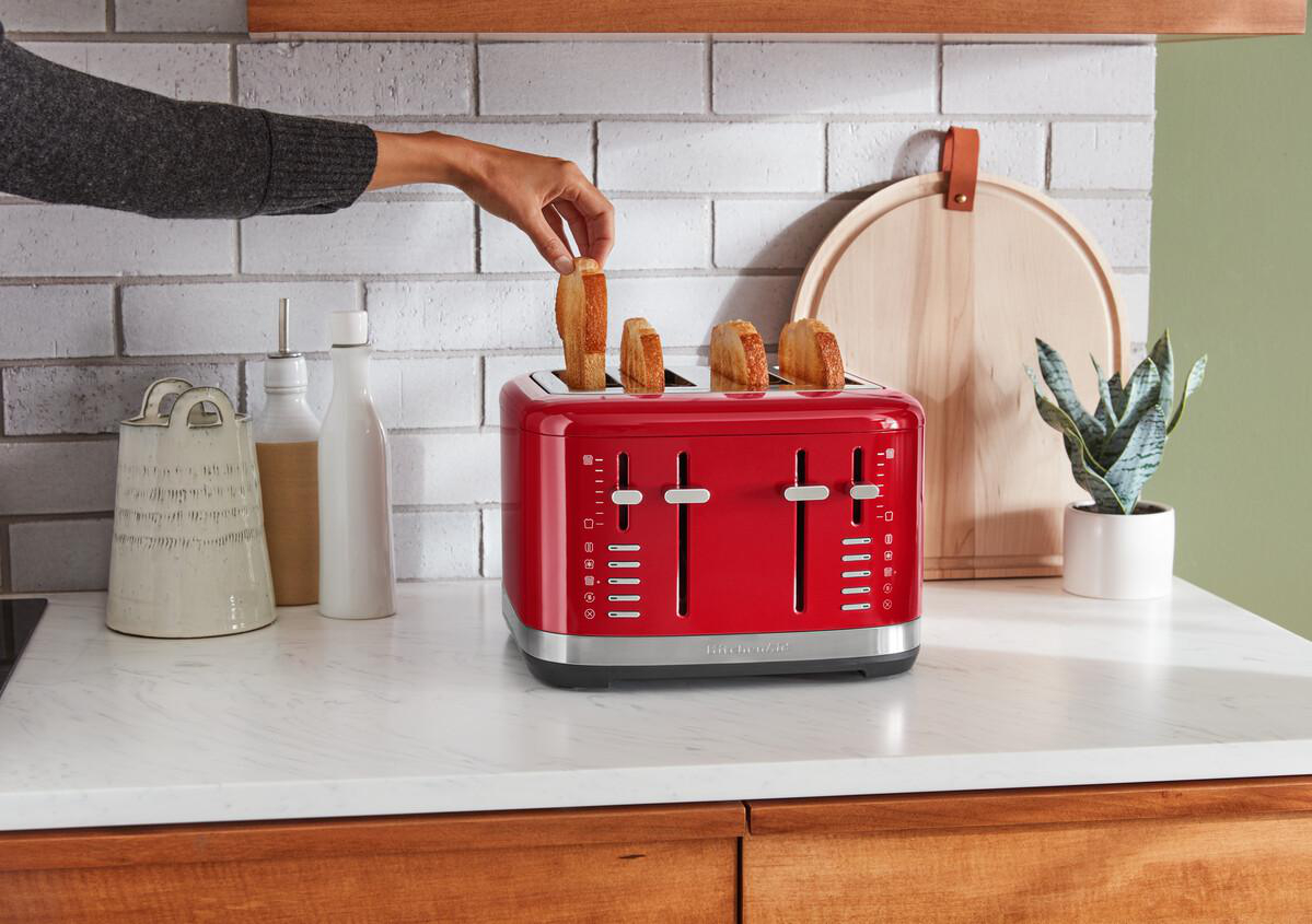 Ein roter Toaster mit vier Schlitzen. Eine Hand legt Brot in die Schlitze, in der Nähe von weißen Flaschen und einer Pflanze.