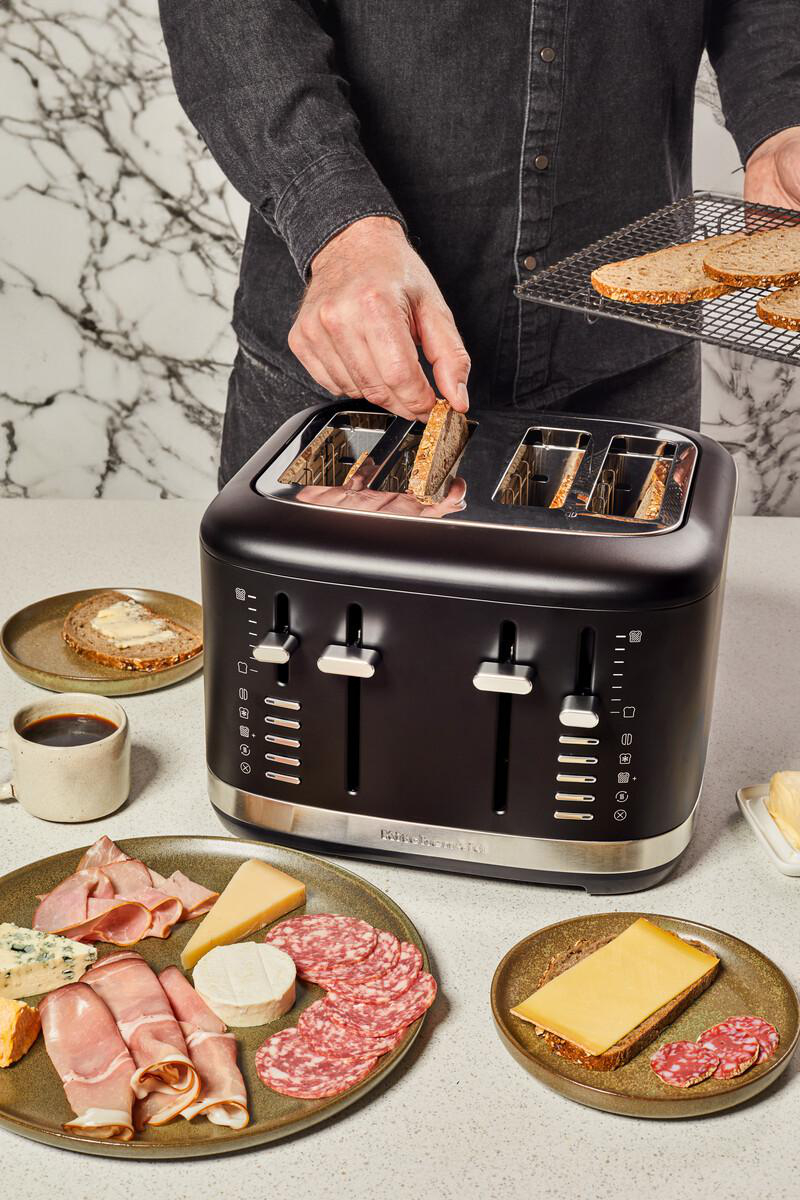 Ein Mann legt Brot in einen schwarzen Toaster. Käse, Fleisch und Butter sind auf Tellern in der Nähe.