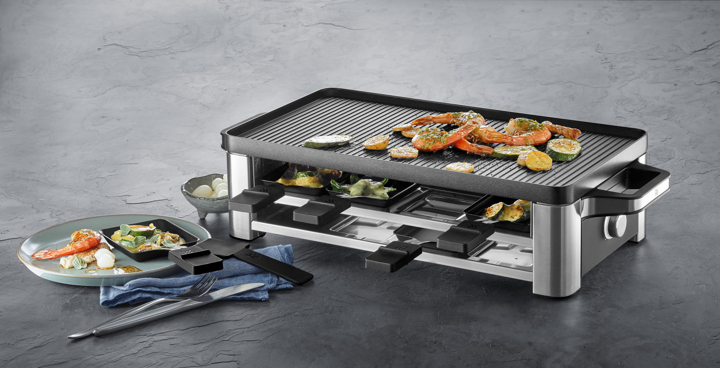 Ein Raclette-Grill mit Gemüse und Garnelen. Es gibt einen Teller mit Essen, Utensilien und eine blaue Serviette.