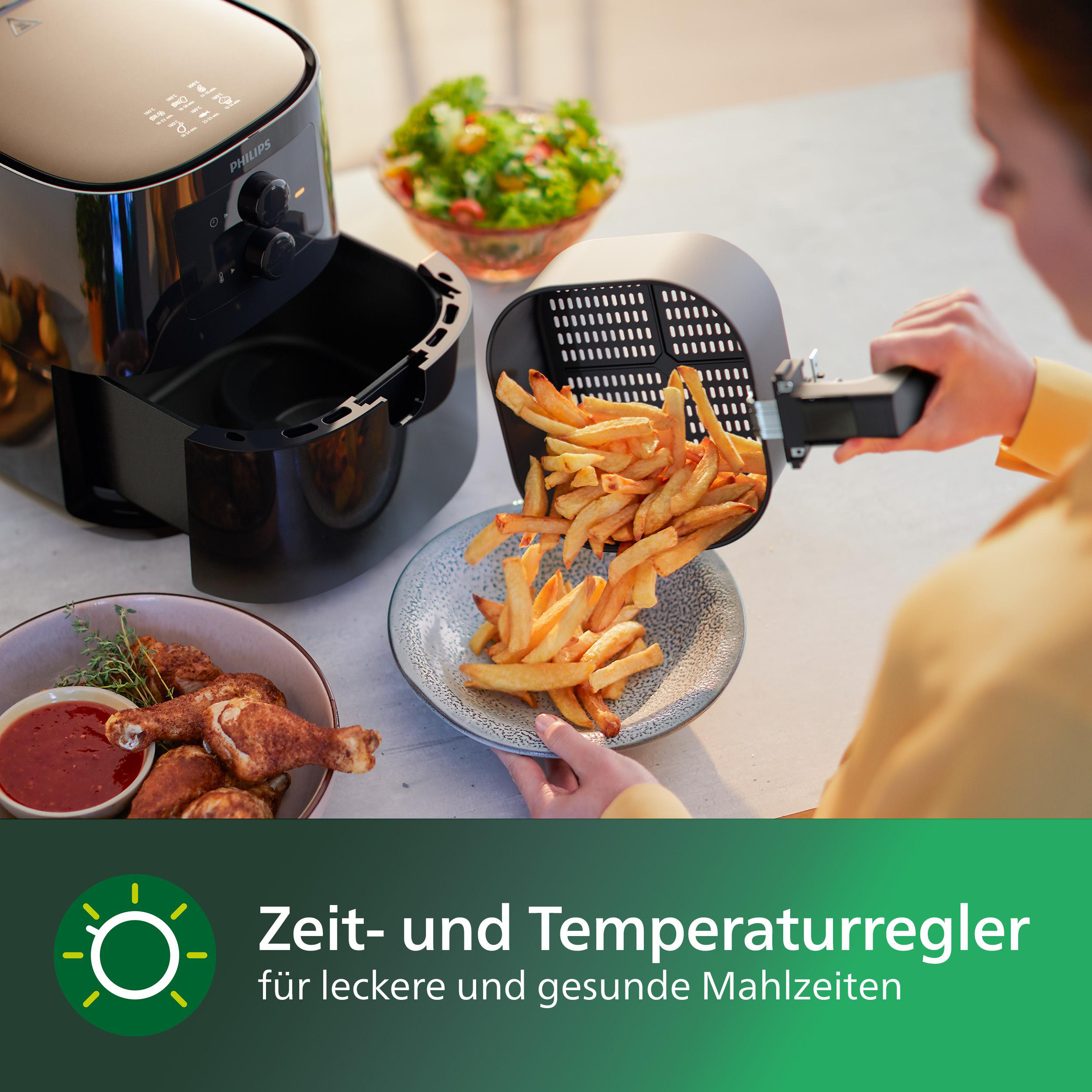 Heißluftfritteuse mit Pommes, die auf einen Teller gelegt werden. Hähnchenkeulen und Salat sind auch vorhanden.
