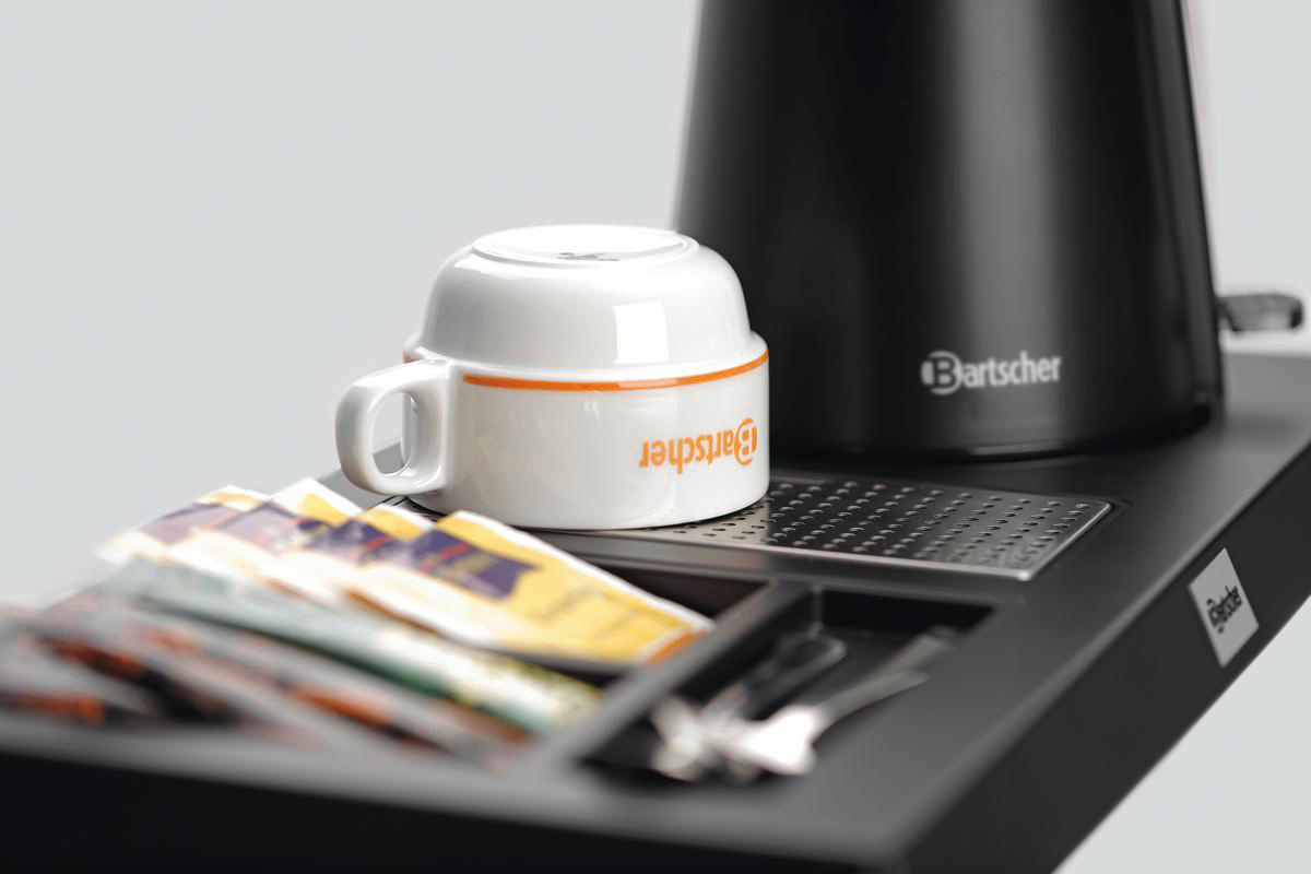 Eine weiße Tasse mit orangefarbenem Rand steht auf einem schwarzen Tablett mit Zuckerpäckchen, einer schwarzen Kaffeemaschine und Besteck.