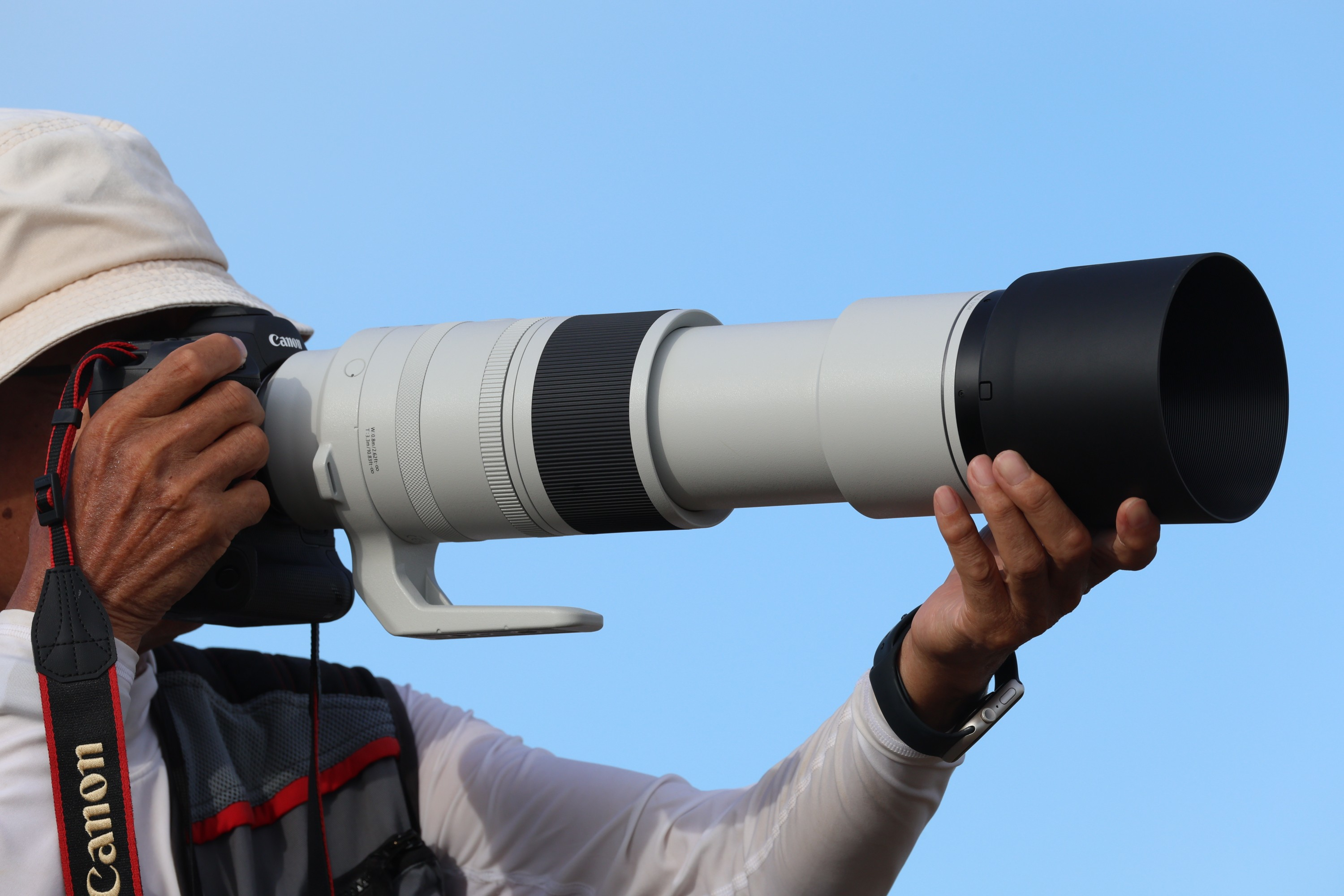 Fotograf hält Canon-Kamera mit langem Objektiv, blauer Himmel im Hintergrund.