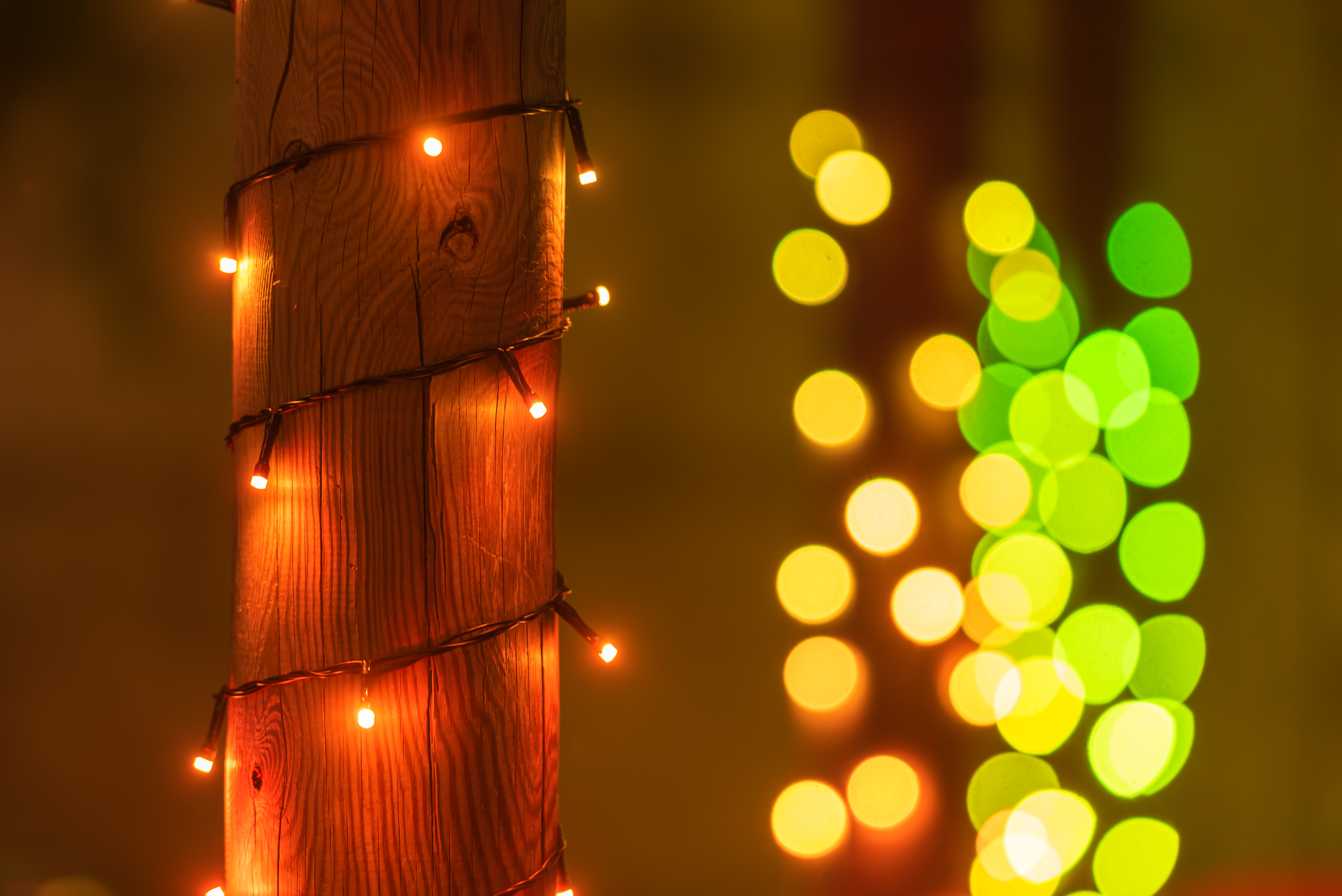 Holzpfahl mit Lichtern umwickelt. Orangefarbene Lichter um den Pfahl, mit unscharfem grünem und gelbem Bokeh.