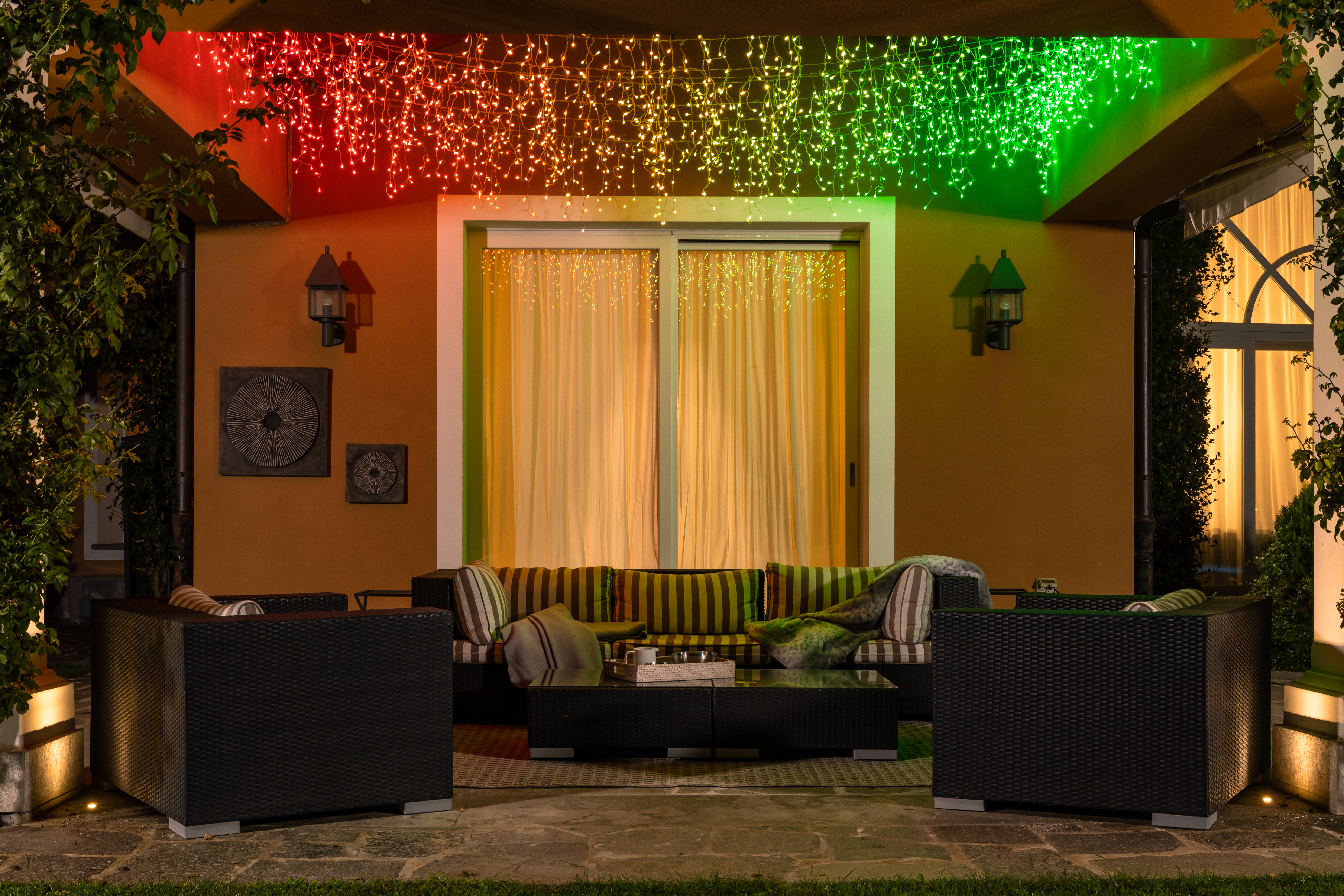 Eine Terrasse mit Sofa, Sesseln und bunten Deckenleuchten. Die Szene hat warme Orange- und Grüntöne.