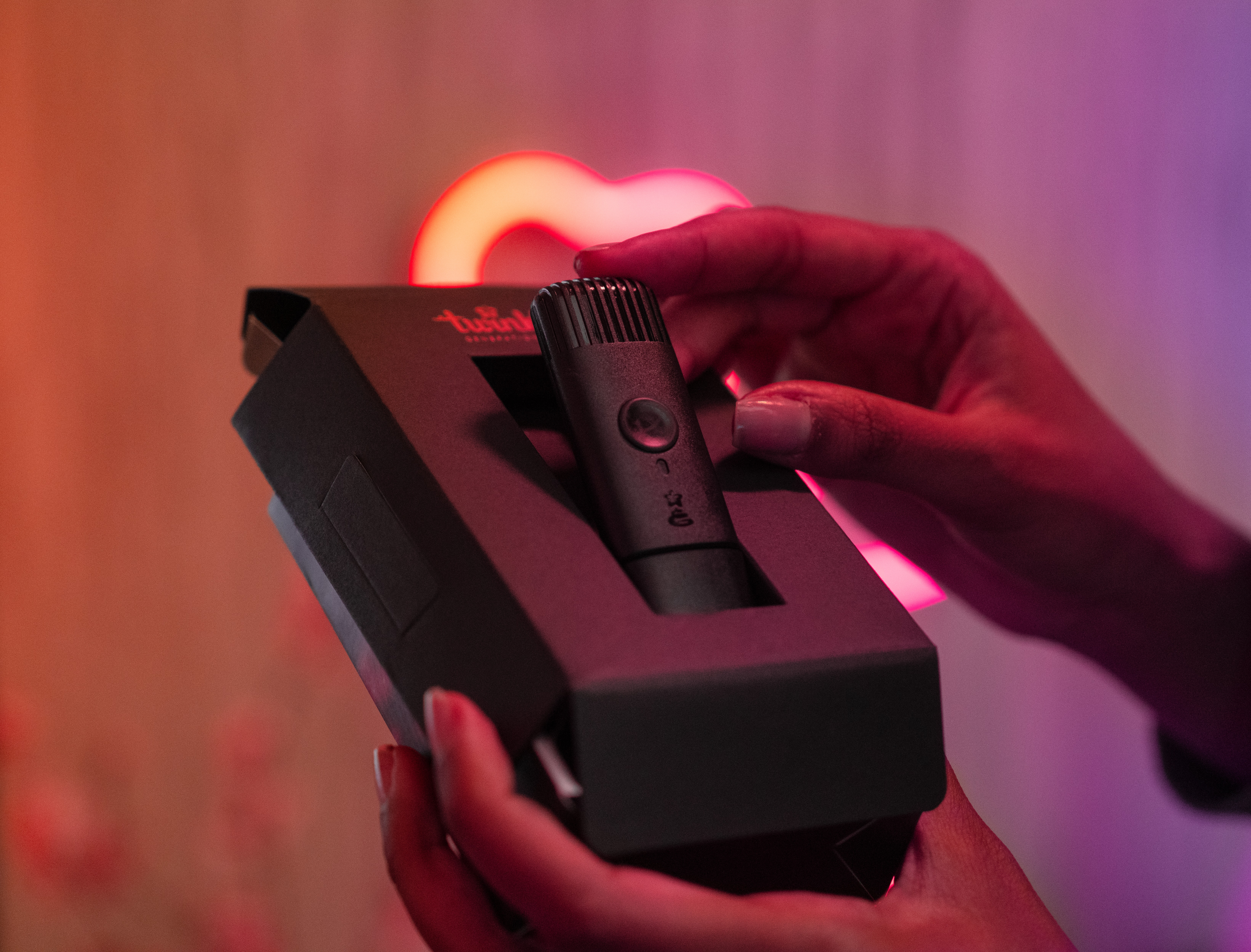 Person hält ein schwarzes Mikrofon in einer schwarzen Box. Im Hintergrund befindet sich ein Neonherz.