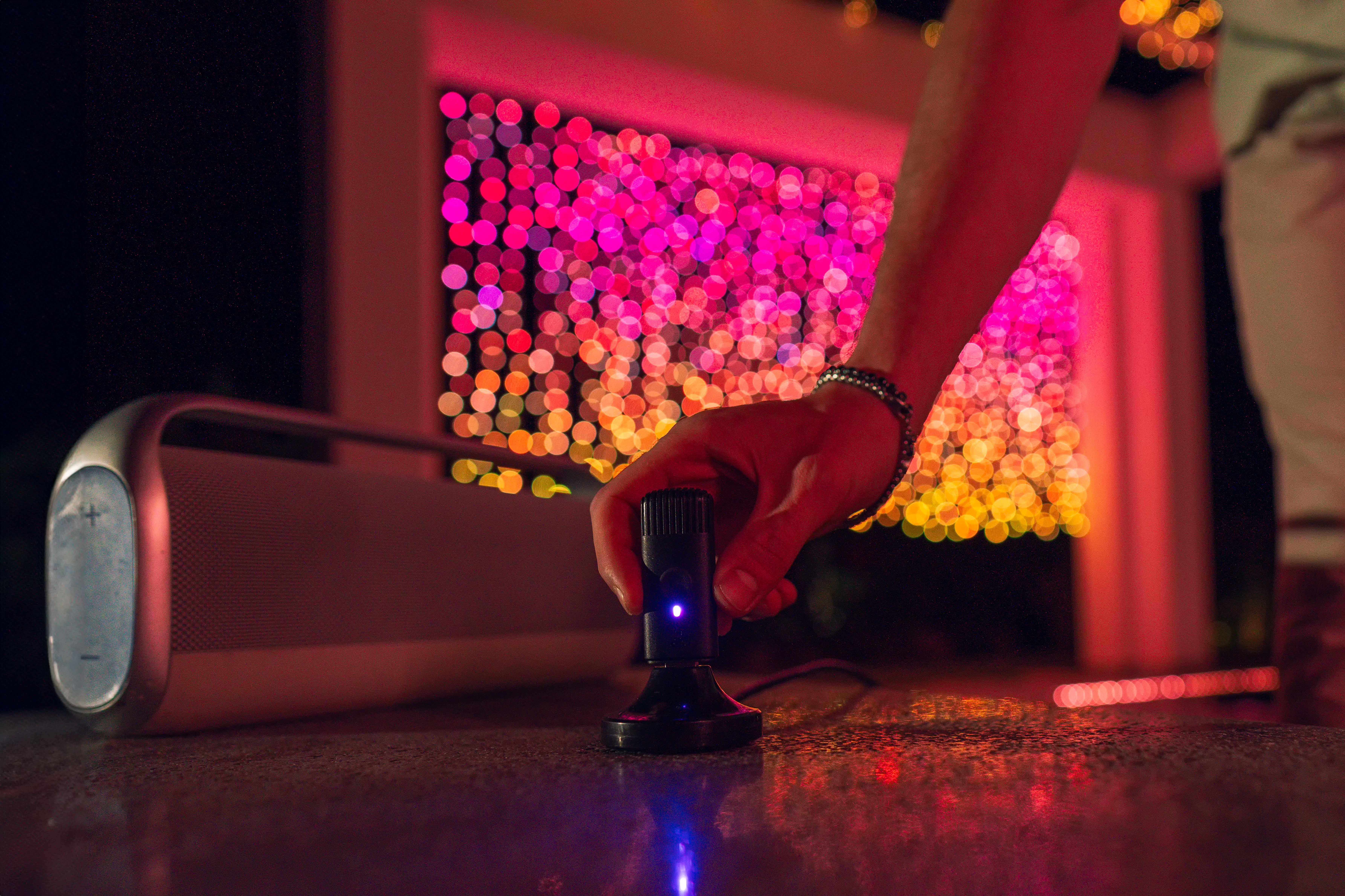 Hand, die ein schwarzes Mikrofon auf einem Tisch hält, neben einem Lautsprecher, Bokeh-Lichter im Hintergrund.