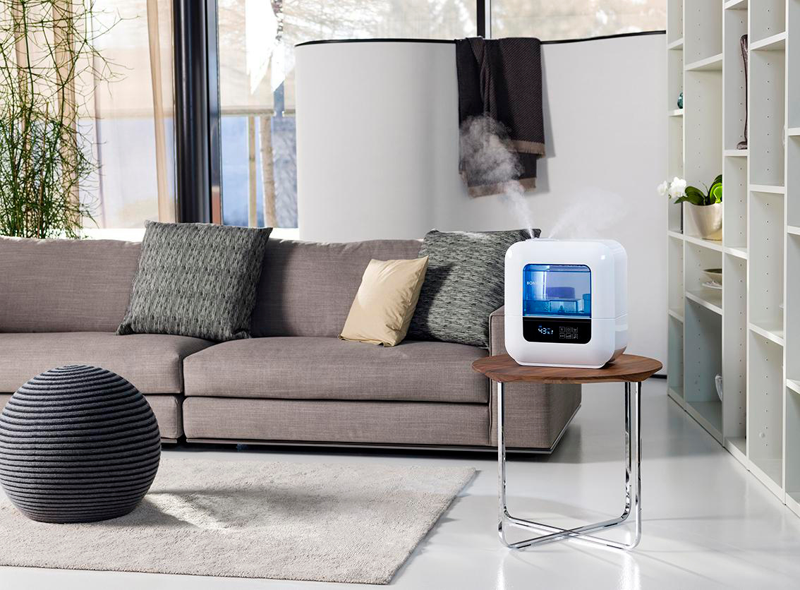 Wohnzimmer mit Sofa, Luftbefeuchter und rundem Pouf. Weiße Regale und ein Fenster sind im Hintergrund.