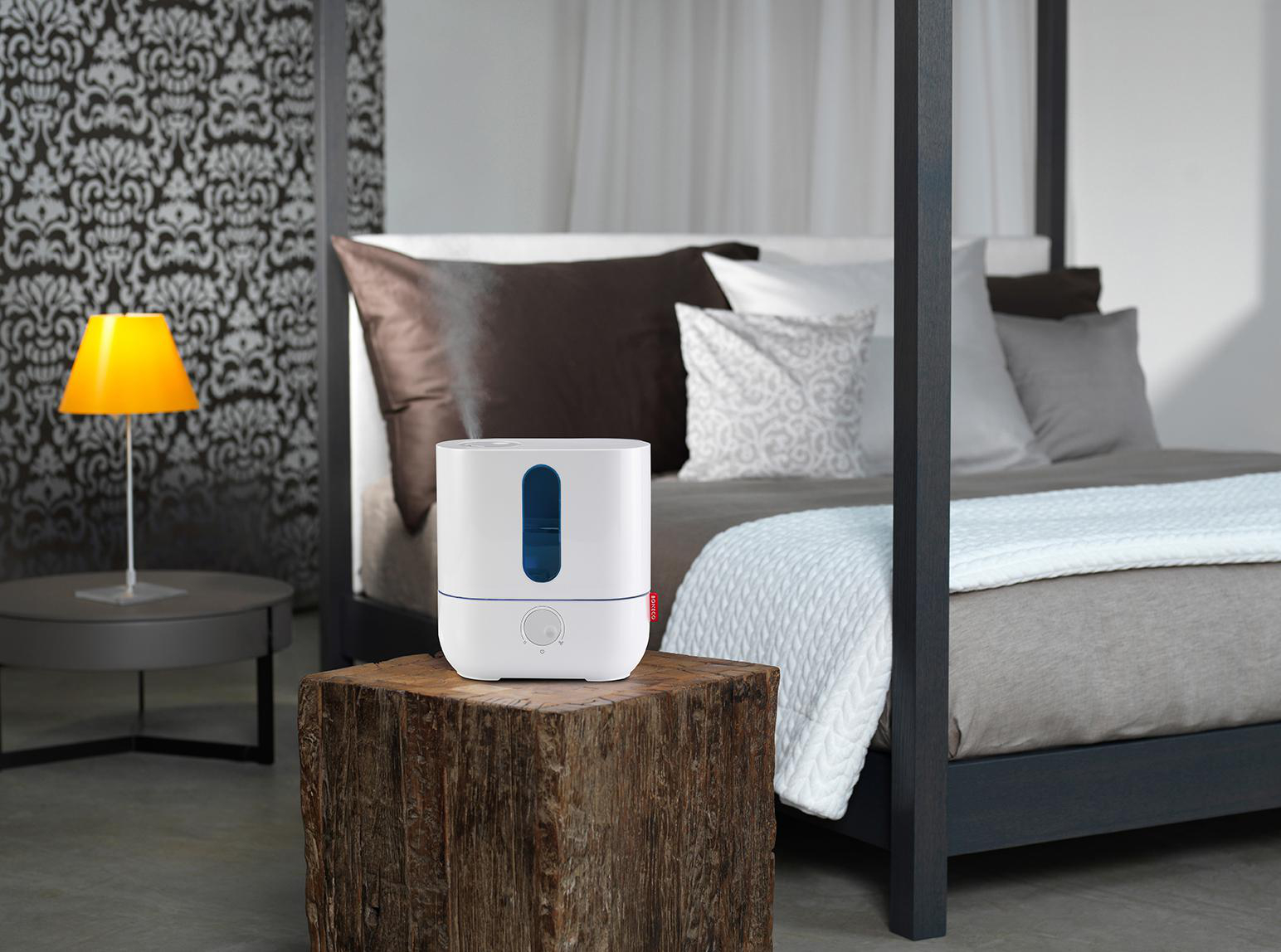 Luftbefeuchter auf einem Holzblock neben einem Bett mit Kissen, Lampe daneben.