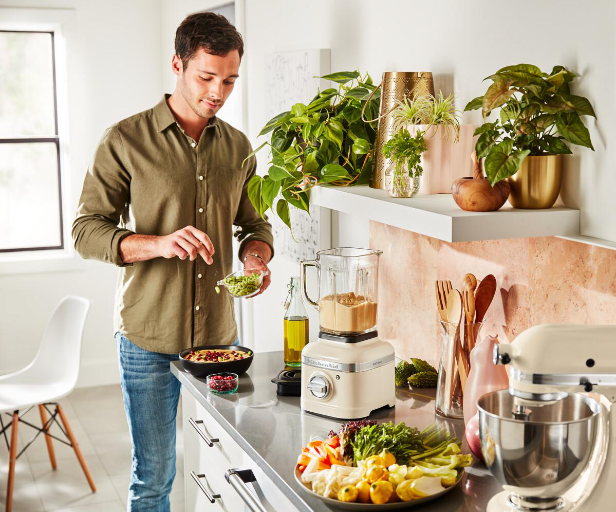 Ein Mann in der Küche bereitet Essen mit einem Mixer und Pflanzen zu.