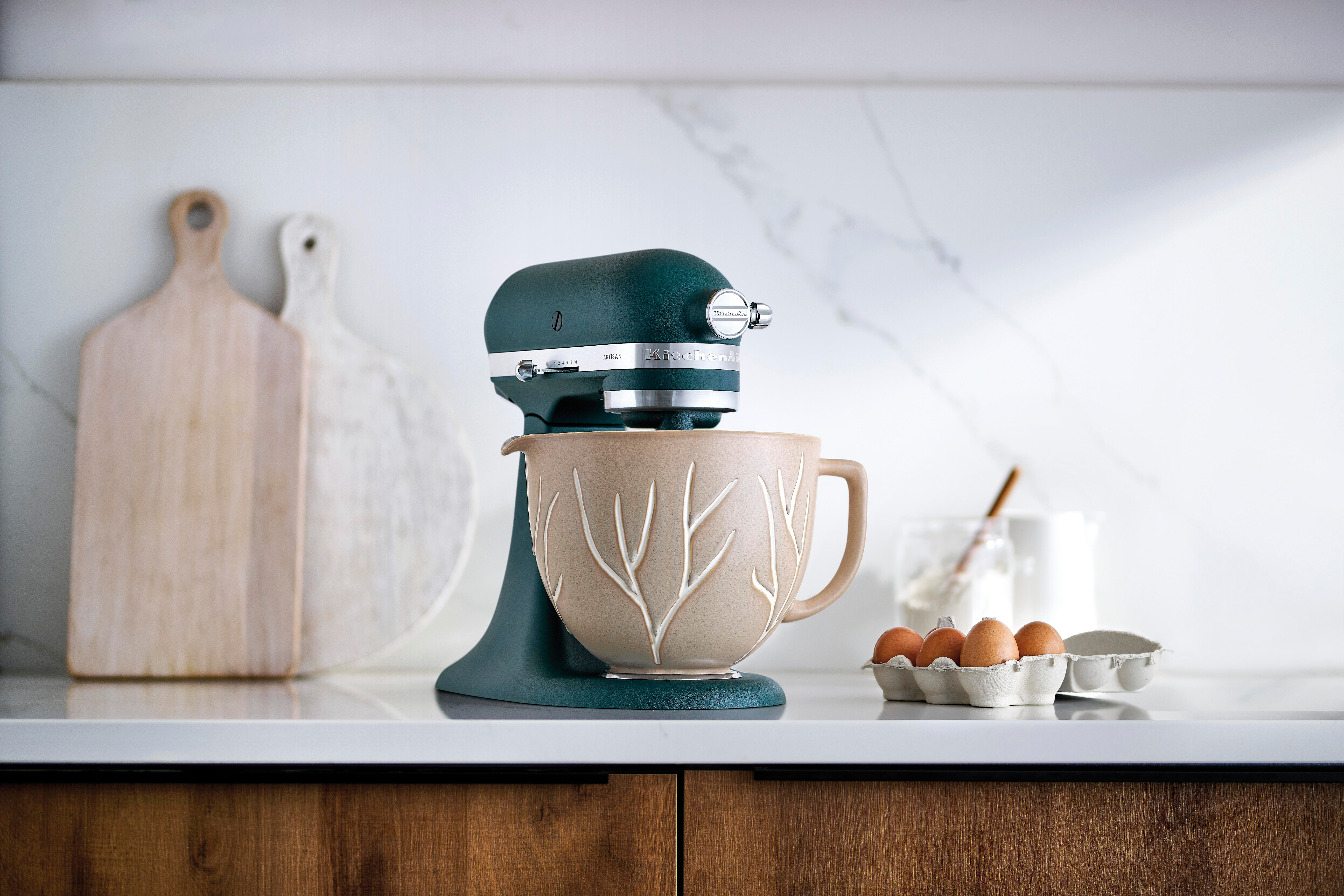 Türkis KitchenAid-Mixer mit Blumenschüssel, Eiern und Schneidebrettern auf einer weißen Arbeitsplatte.