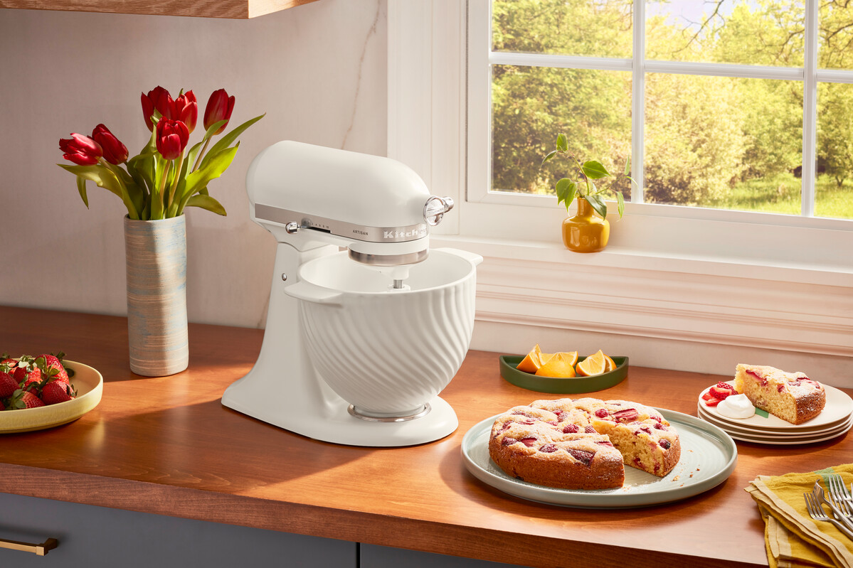 Ein weißer KitchenAid-Mixer mit Kuchen, Obst und Blumen auf einer hölzernen Arbeitsplatte in der Nähe eines Fensters.