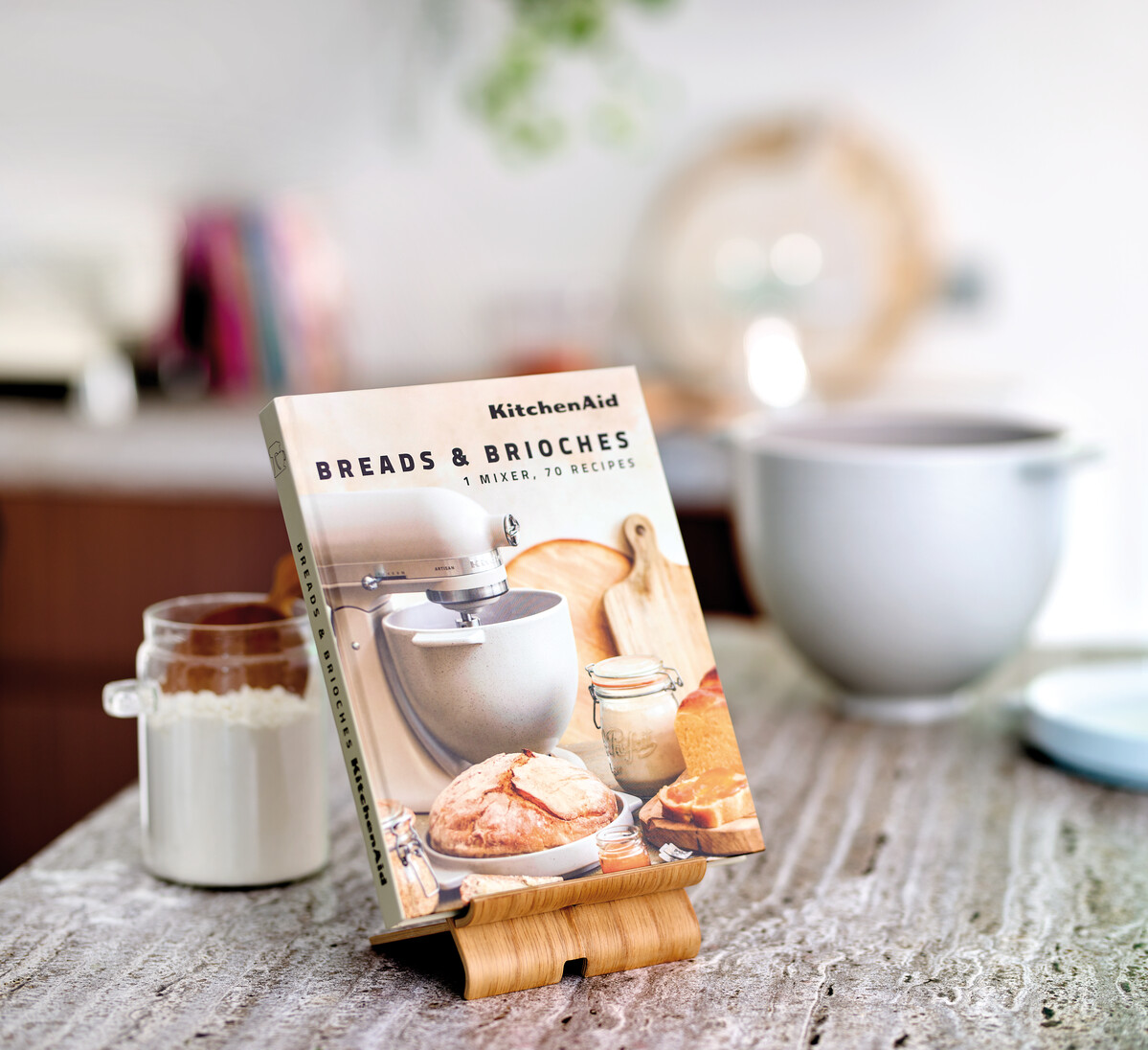 Ein Kochbuch mit dem Titel 'Breads & Brioches' liegt auf einem Holzständer. Küchenutensilien und ein Glas Mehl sind im Hintergrund.