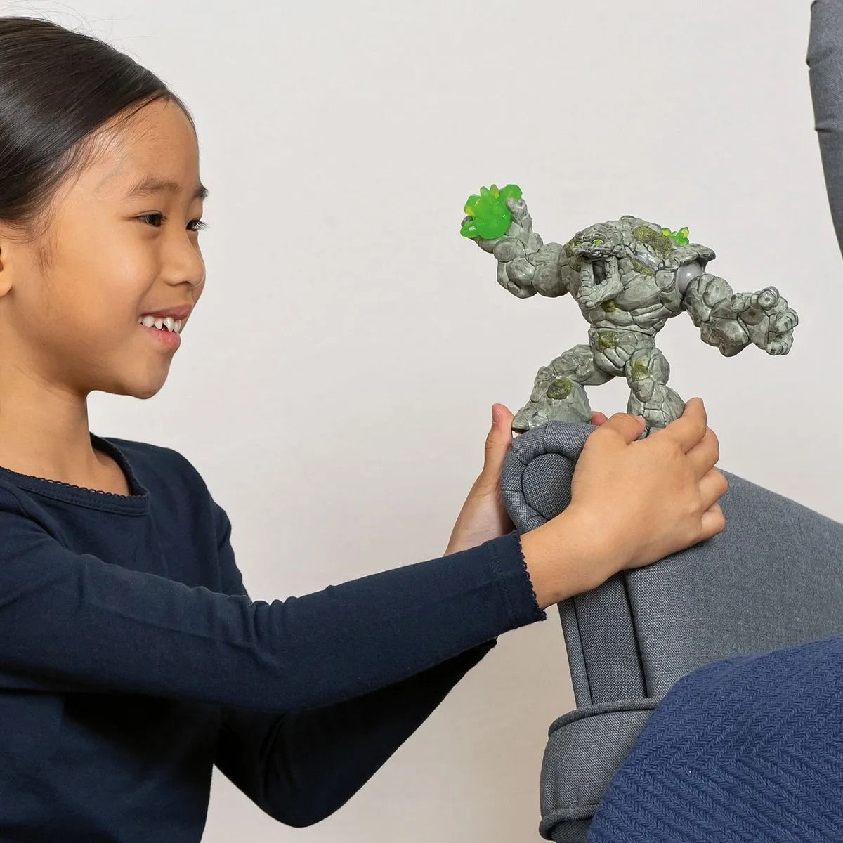 Ein Mädchen lächelt und hält ein Spielzeugmonster. Sie ist in einem Raum mit einem grauen Stuhl.