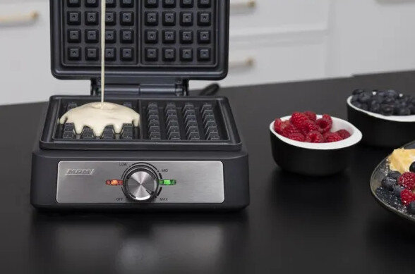 Gofrownica z wylewanym ciastem. Czarna powierzchnia, obok małe miski z jagodami.