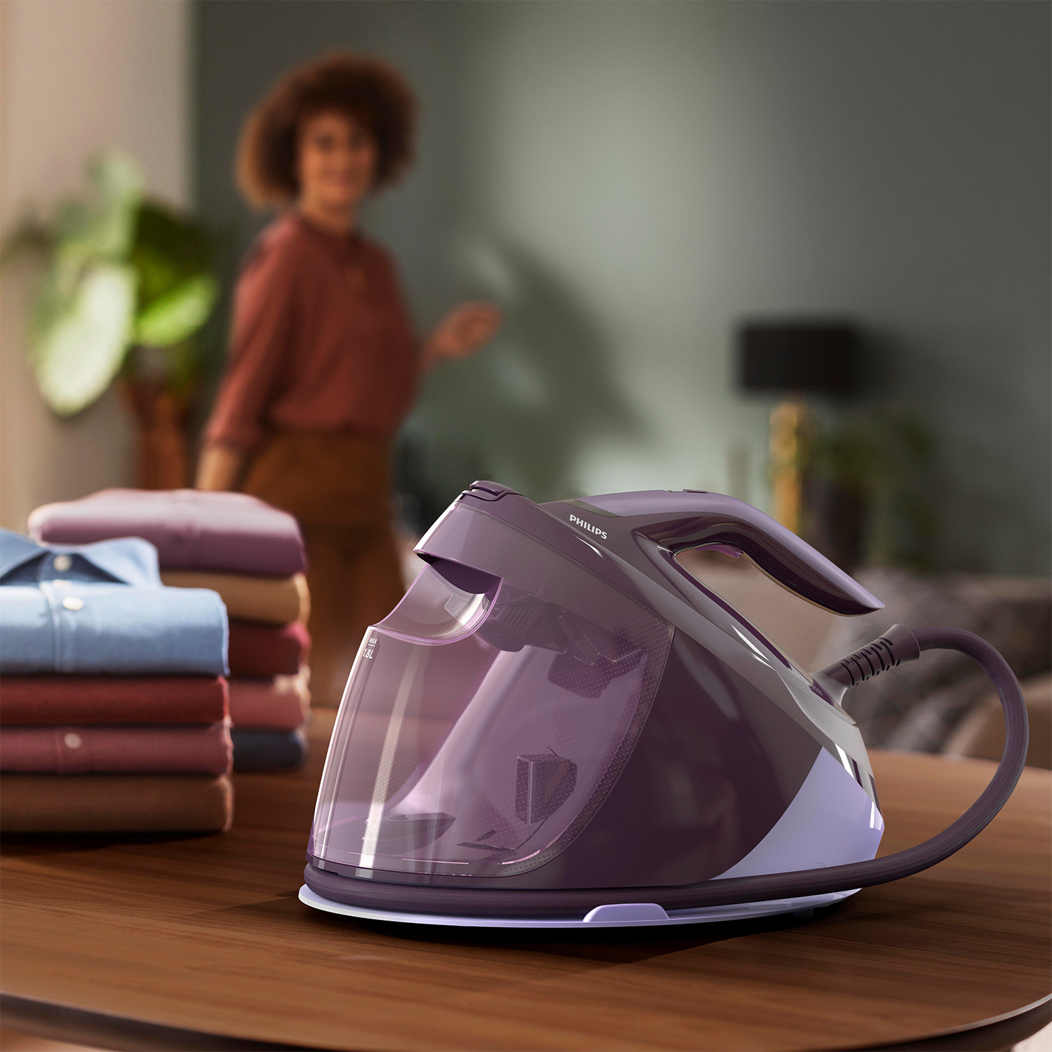 Allestimento stiratura: ferro viola su tavolo, vestiti impilati, donna sfocata sullo sfondo.