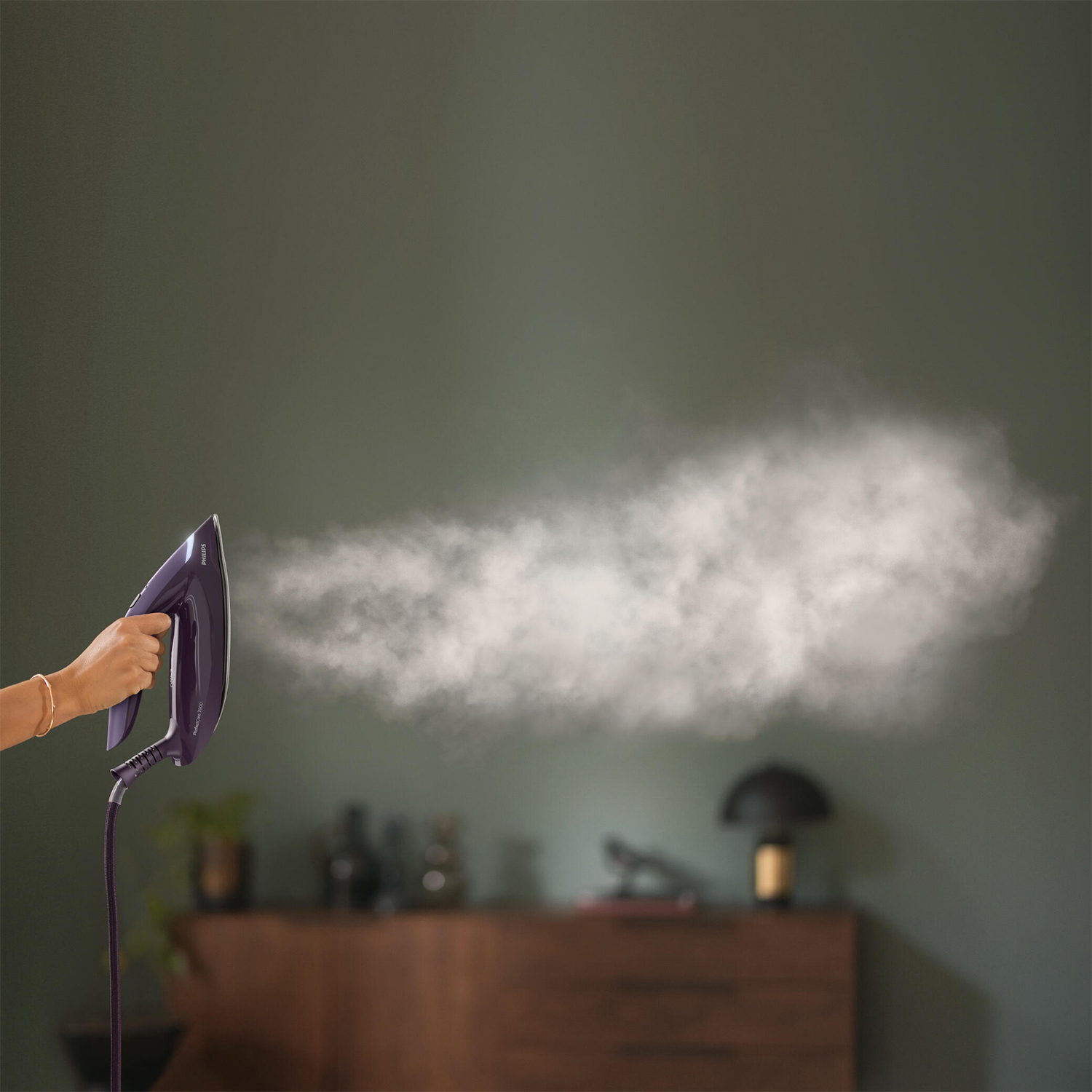 Mano che tiene un ferro da stiro viola, esce vapore. Lo sfondo è un muro verde.