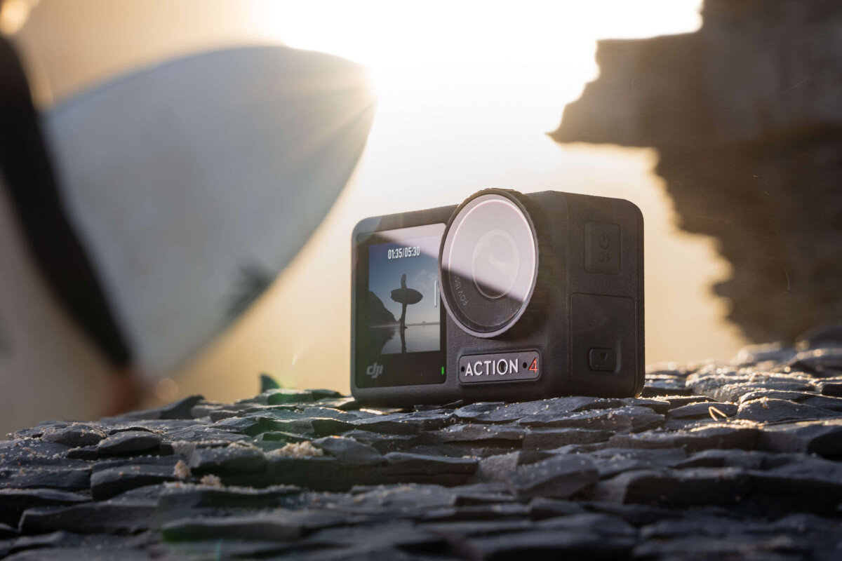 Czarna kamera sportowa na skale, z surferem w tle i świecącym słońcem.