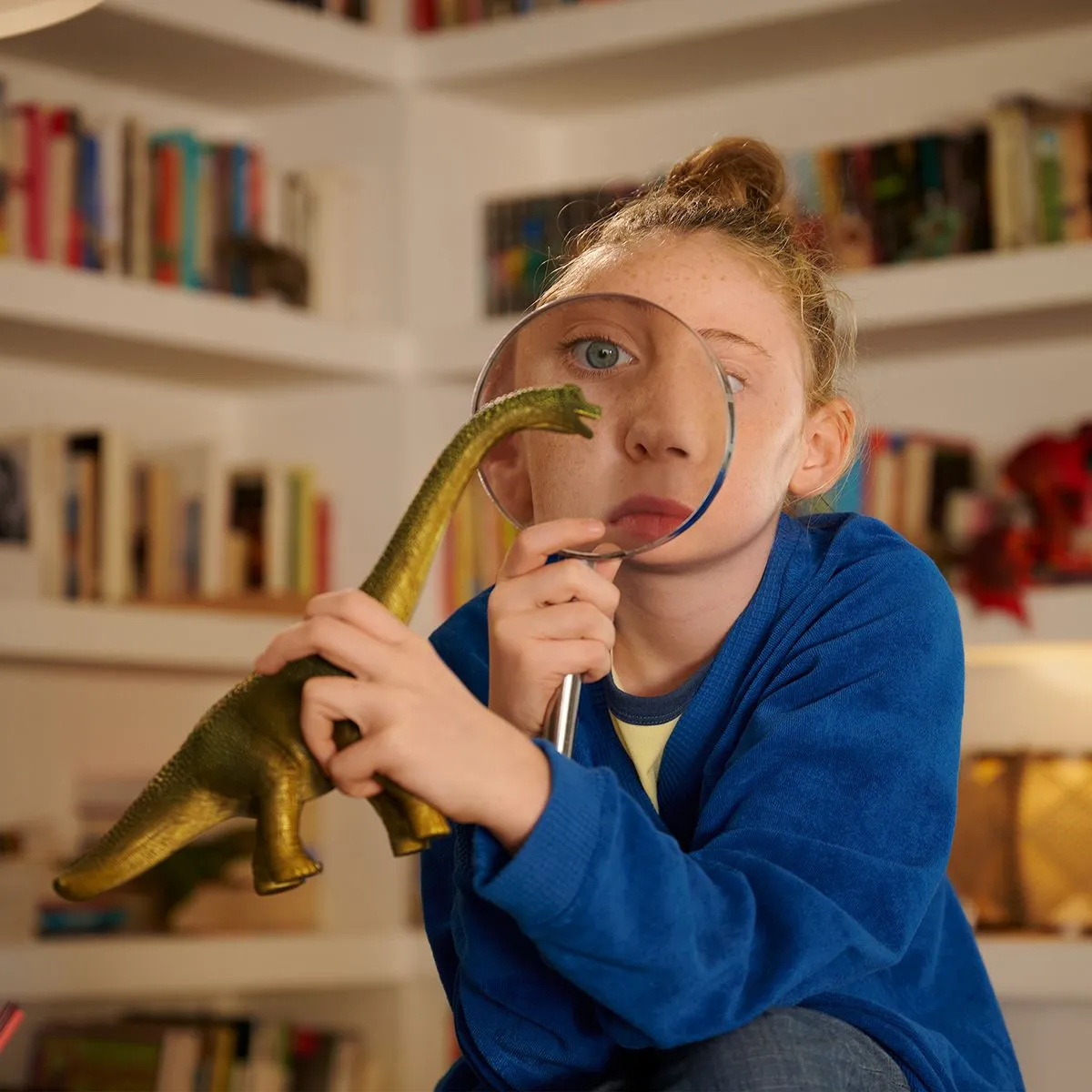 Ein Mädchen schaut durch eine Lupe auf ein Spielzeug-Dinosaurier, Bücher im Hintergrund.