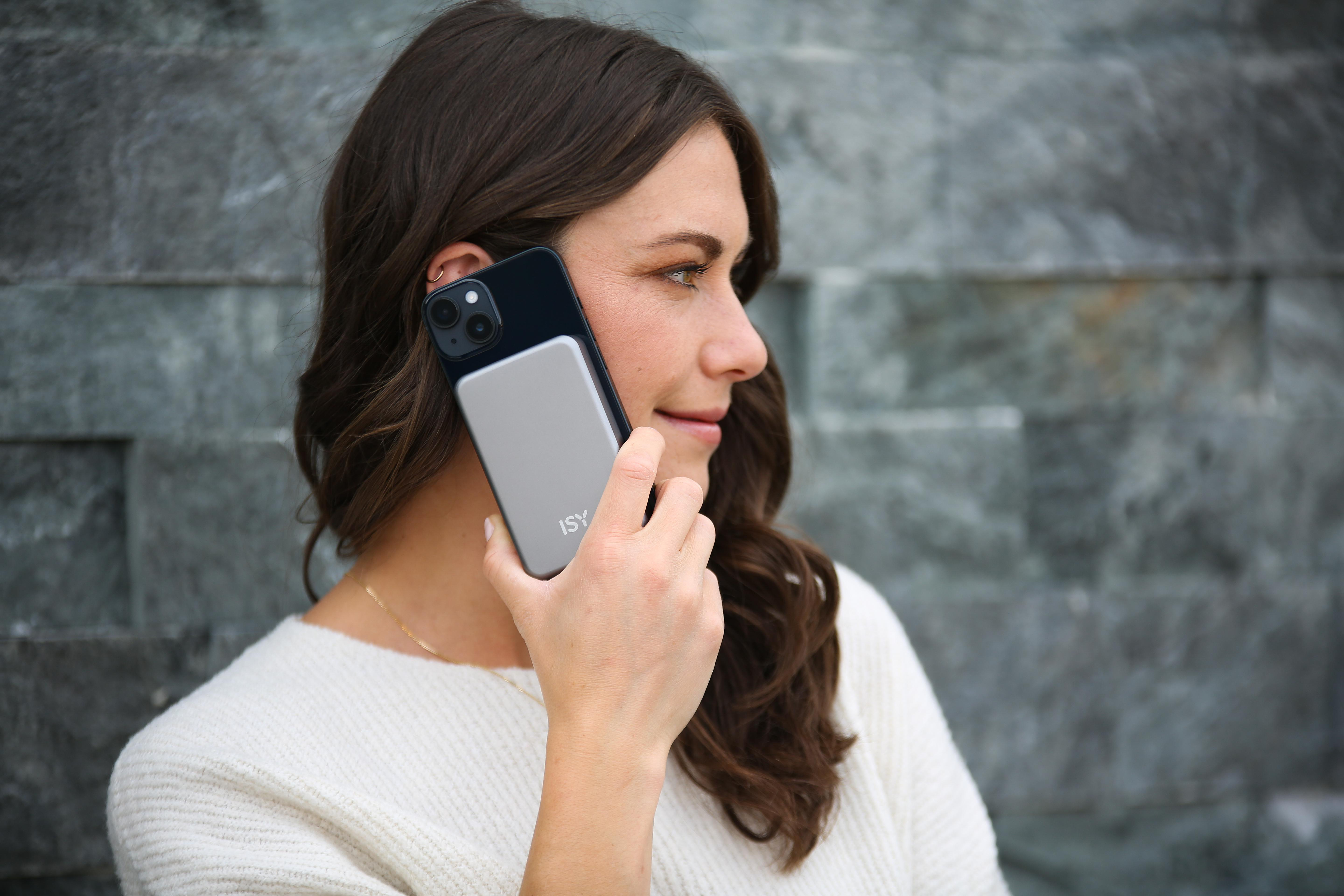 Frau hält ein schwarzes Telefon mit einem silbernen Akkupack. Sie lächelt, vor einem grauen Steinmauer-Hintergrund.