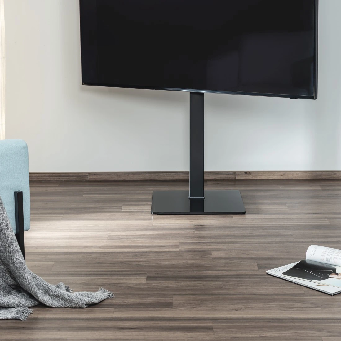 Una TV nera montata su un supporto nero su un pavimento in legno. Un muro bianco sullo sfondo.