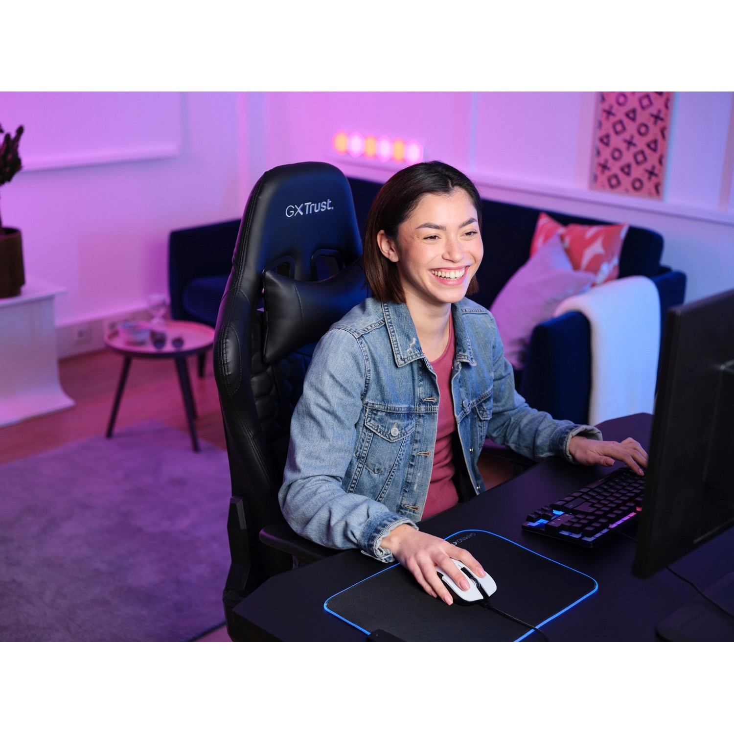 A smiling woman using a computer with a black desk, black chair, and blue and pink lighting.
