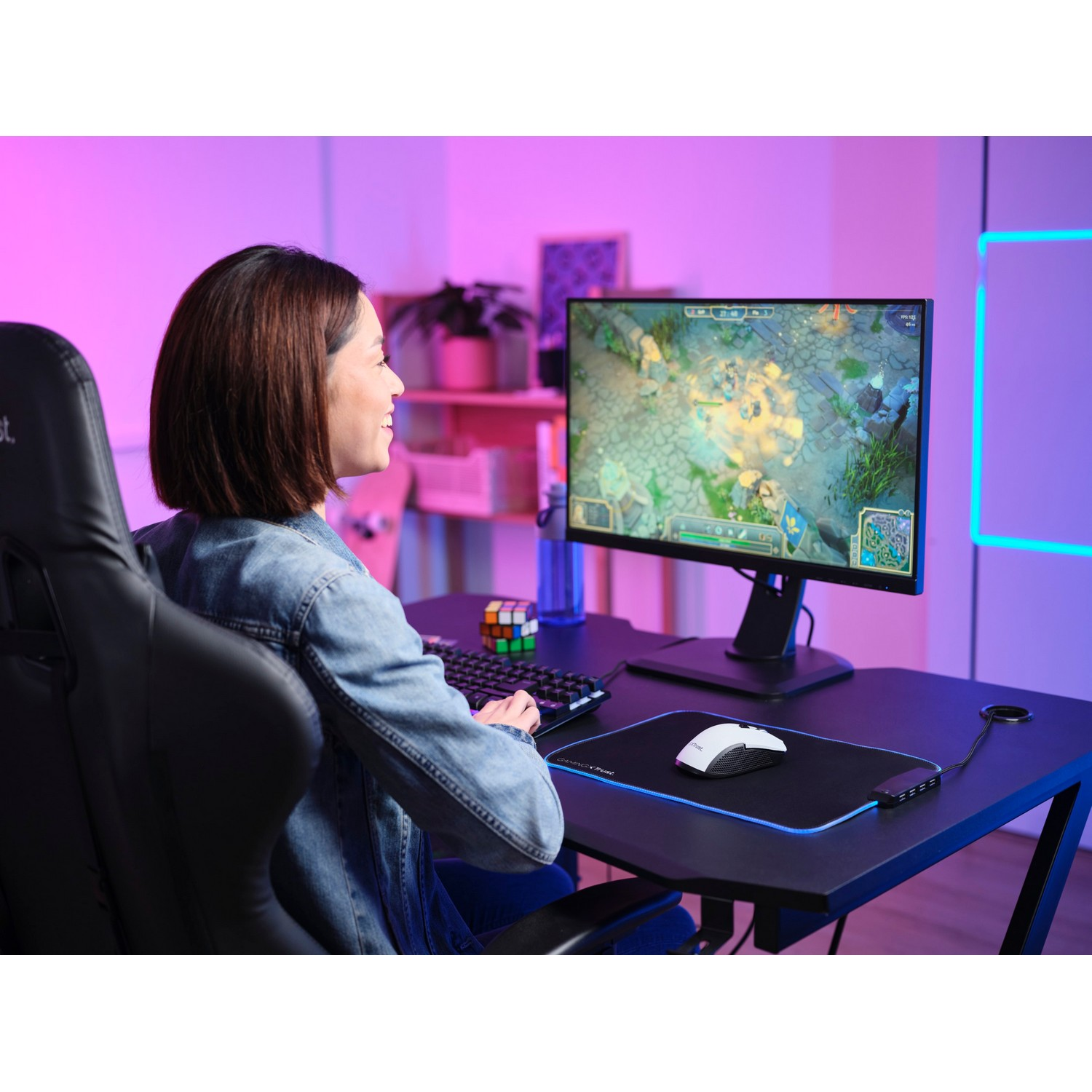 A woman sits at a desk, using a computer. Monitor showing a game, colorful lights.