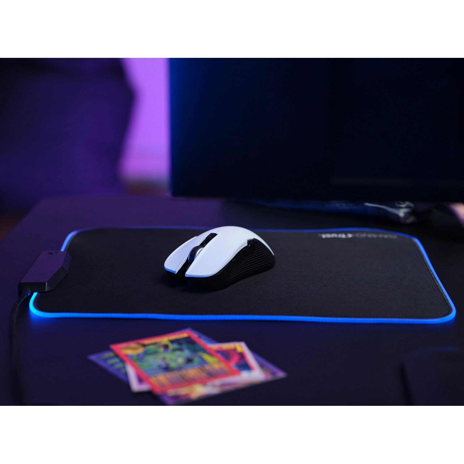 Gaming setup with white mouse, black mousepad with blue light, and cards. Monitor in background.