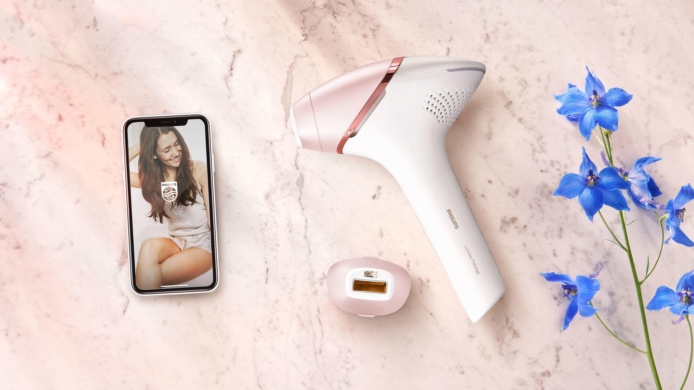 IPL-Haarentfernungsgerät, Telefon mit App-Bildschirm und blaue Blumen auf rosa Oberfläche.