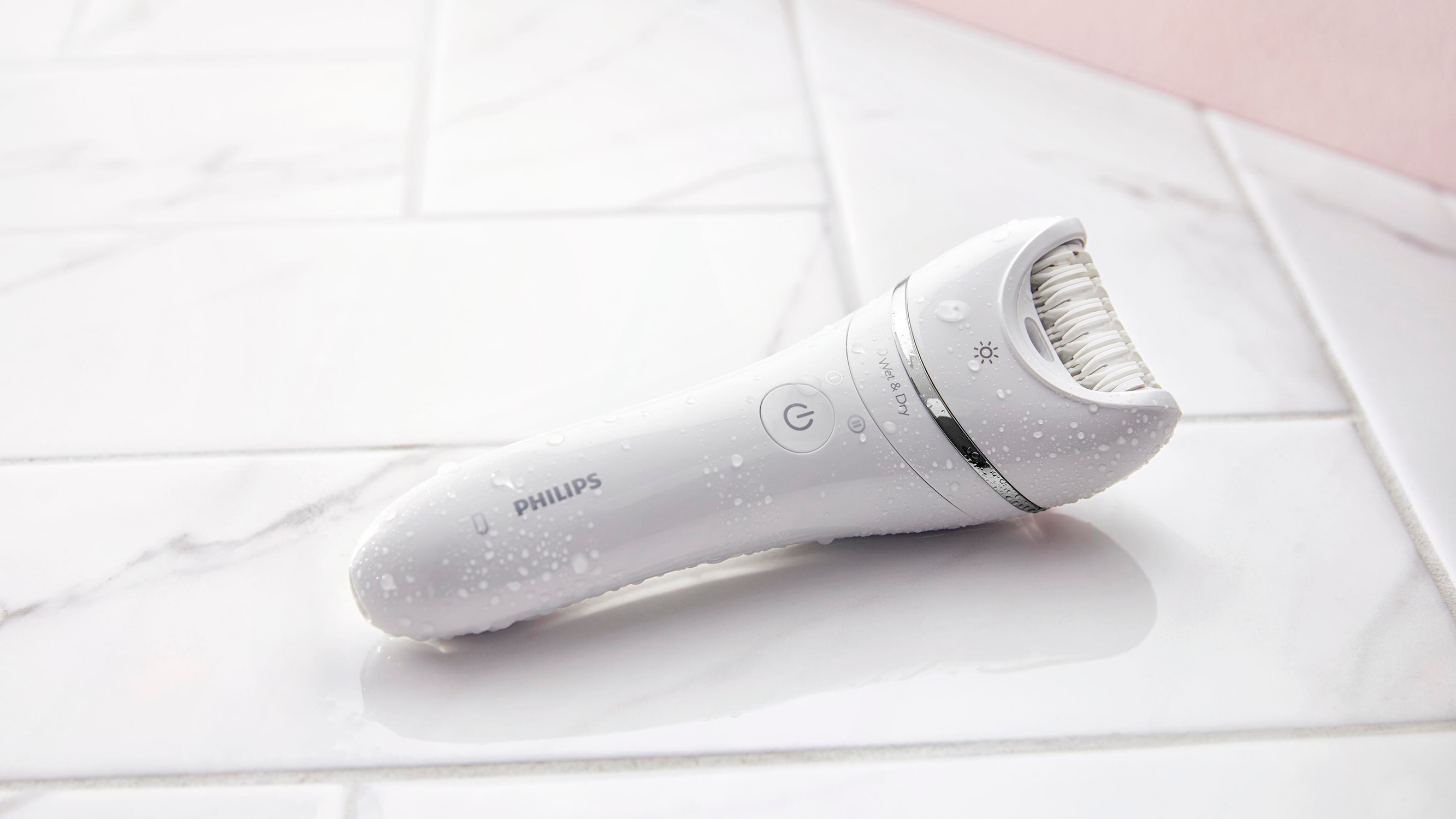 Een witte Philips epilator op een wit betegeld oppervlak, nat met waterdruppels.