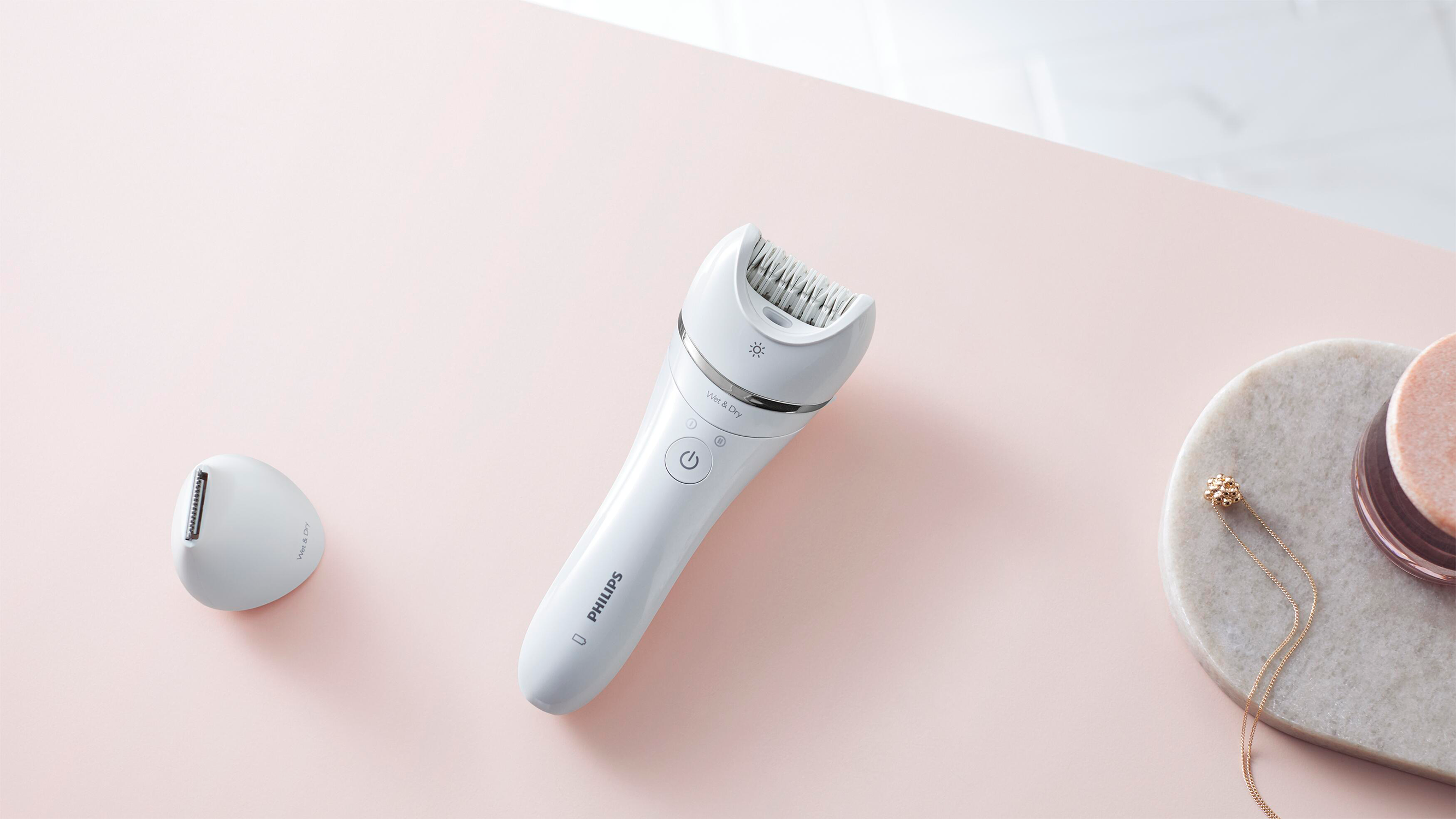 Witte elektrische epilator en losse kop op roze oppervlak. Gouden ketting op marmer.