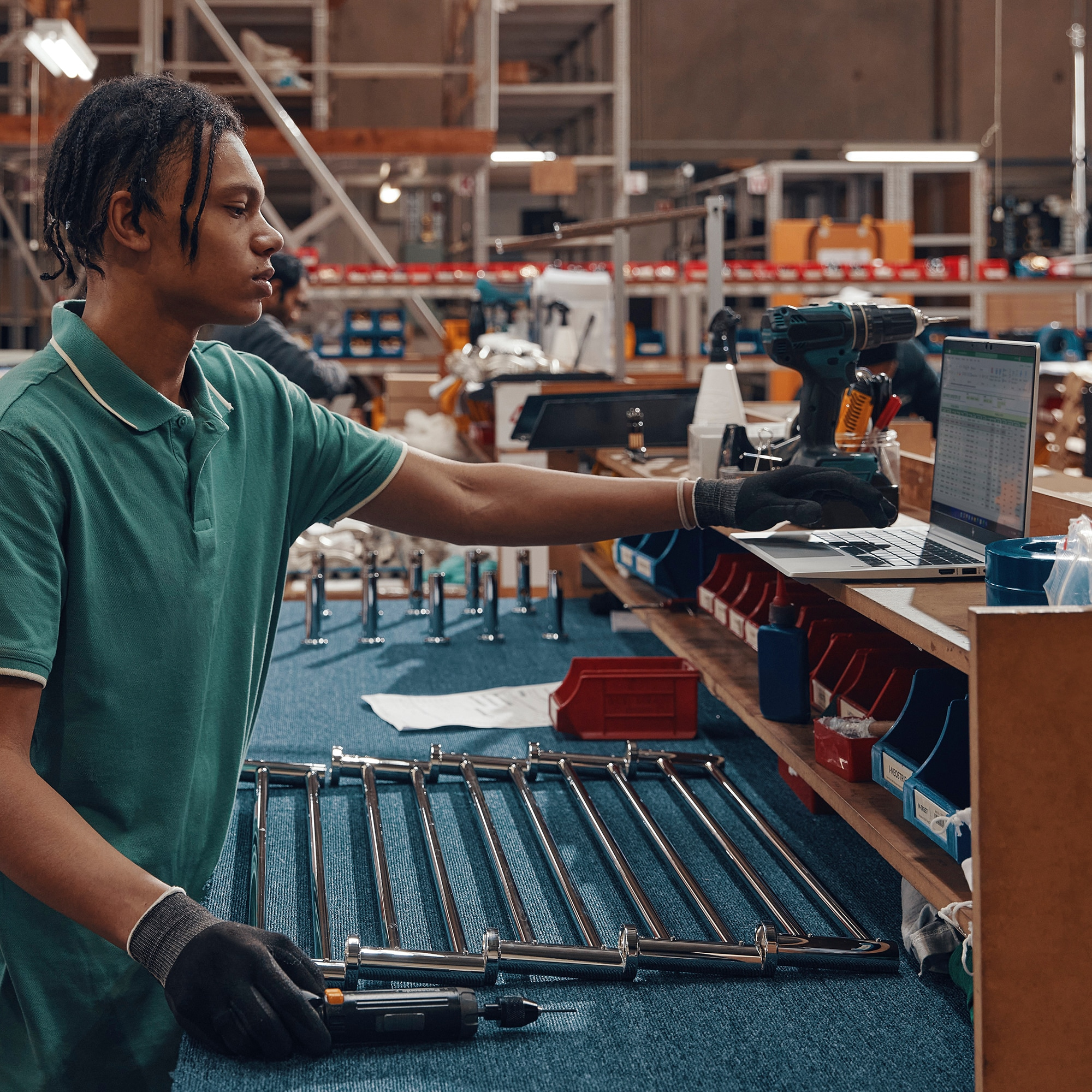 Ein Mann in grünem Hemd arbeitet mit Werkzeugen und einem Laptop an einer Metallstruktur.