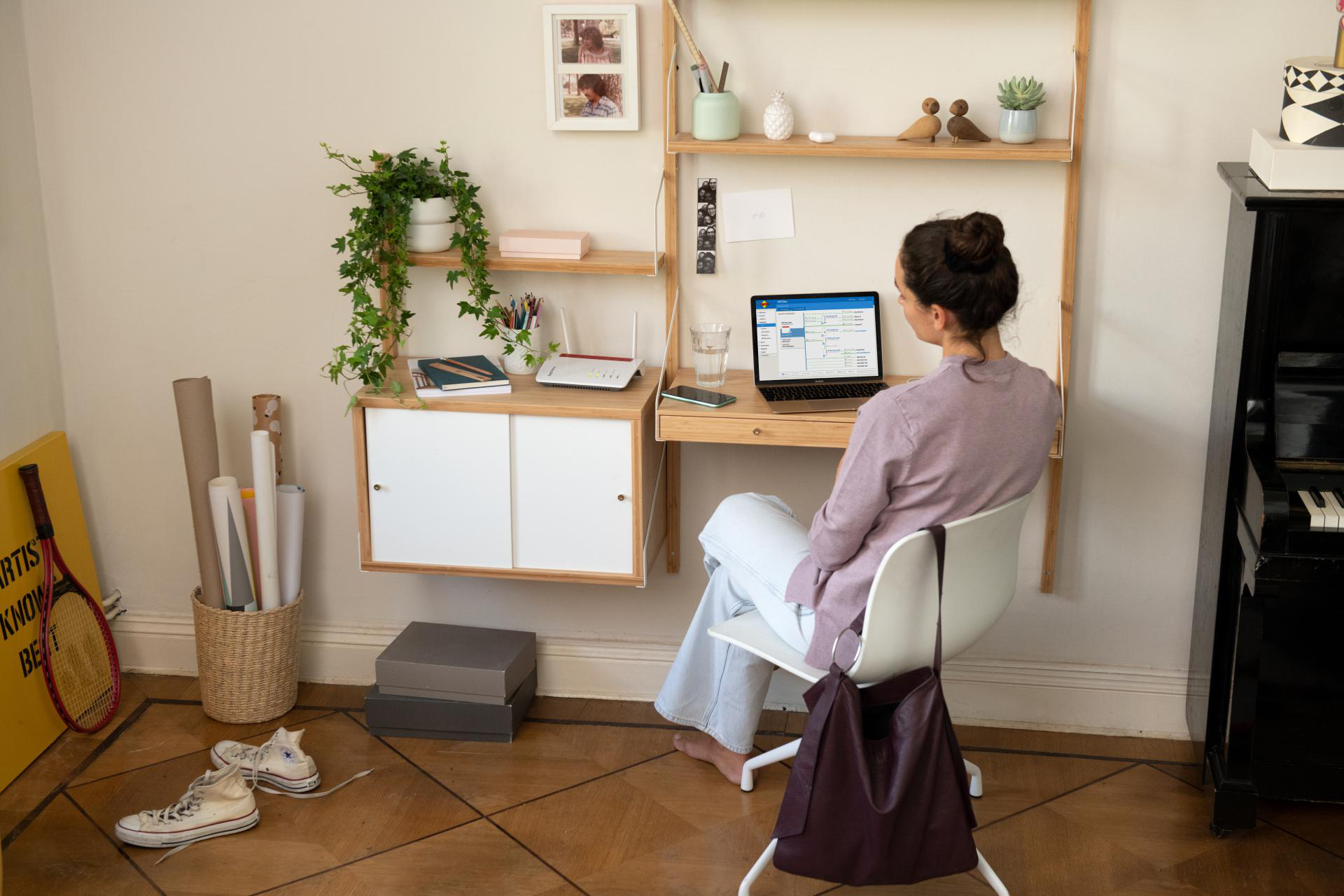 Frau am Schreibtisch, Laptop offen, Pflanzen, Bücher und weiße Wand.