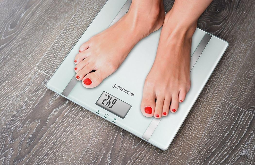 Nő lábai egy fehér és ezüst mérlegen, 64,2 súlyt mutatva. Piros körömlakk látható.