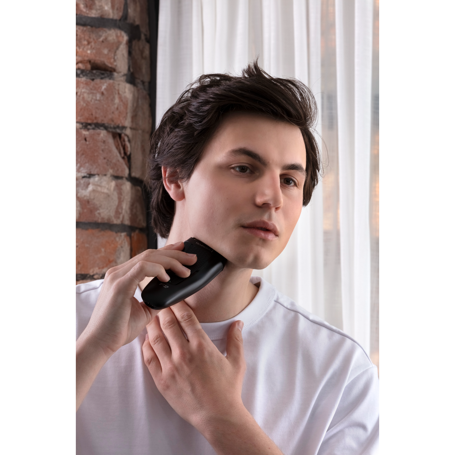 Man shaving his face with a black electric shaver. White t-shirt, brick wall background.