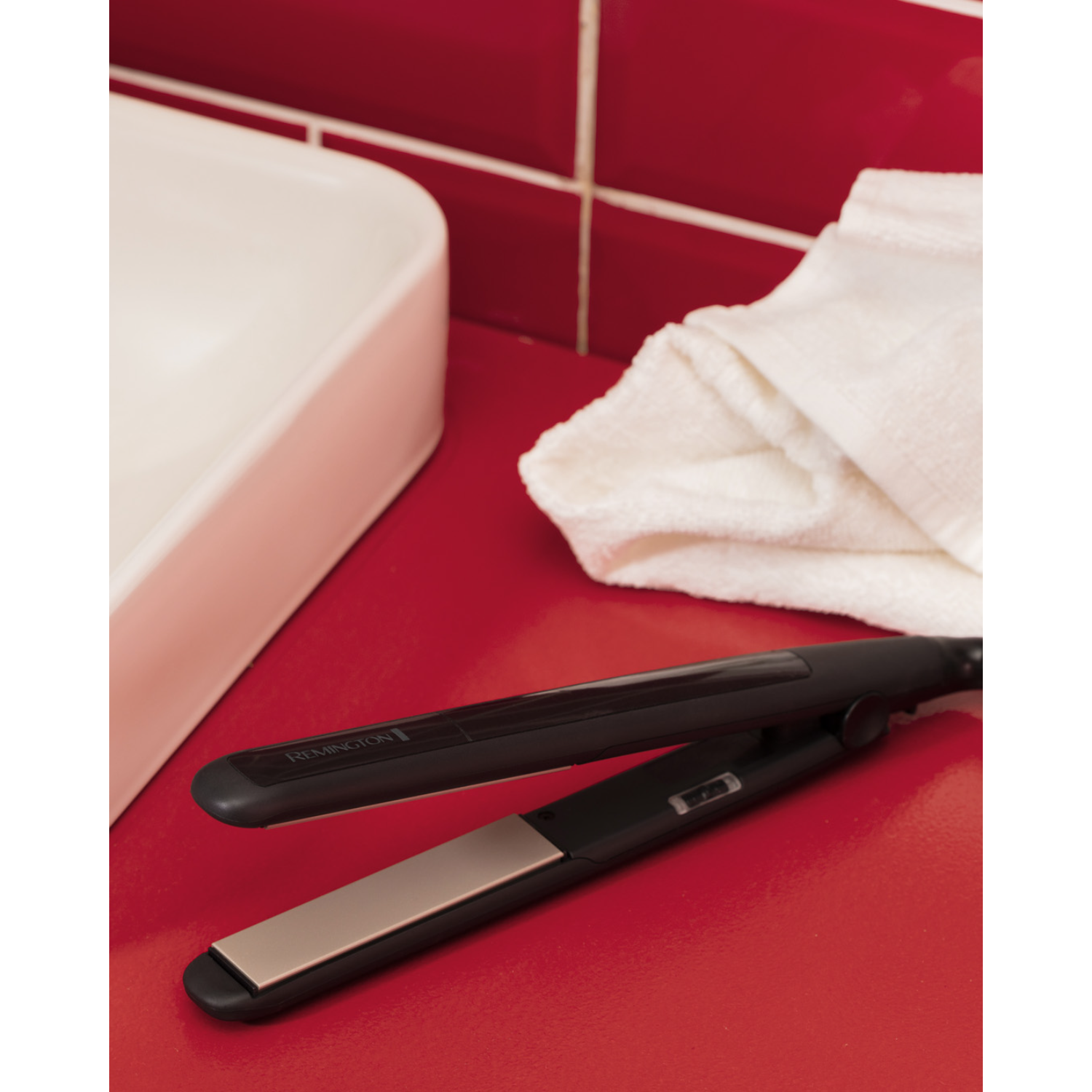 Hair straightener on red surface. White sink and towel in the background. Black and gold color.