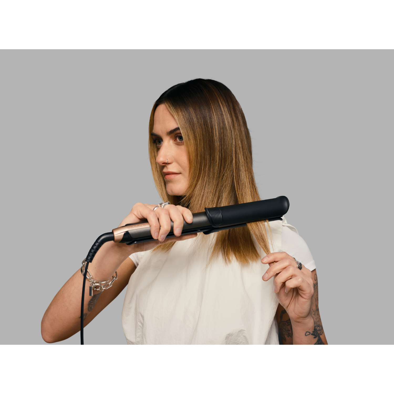 A woman is straightening hair with a black and gold flat iron. Gray background.
