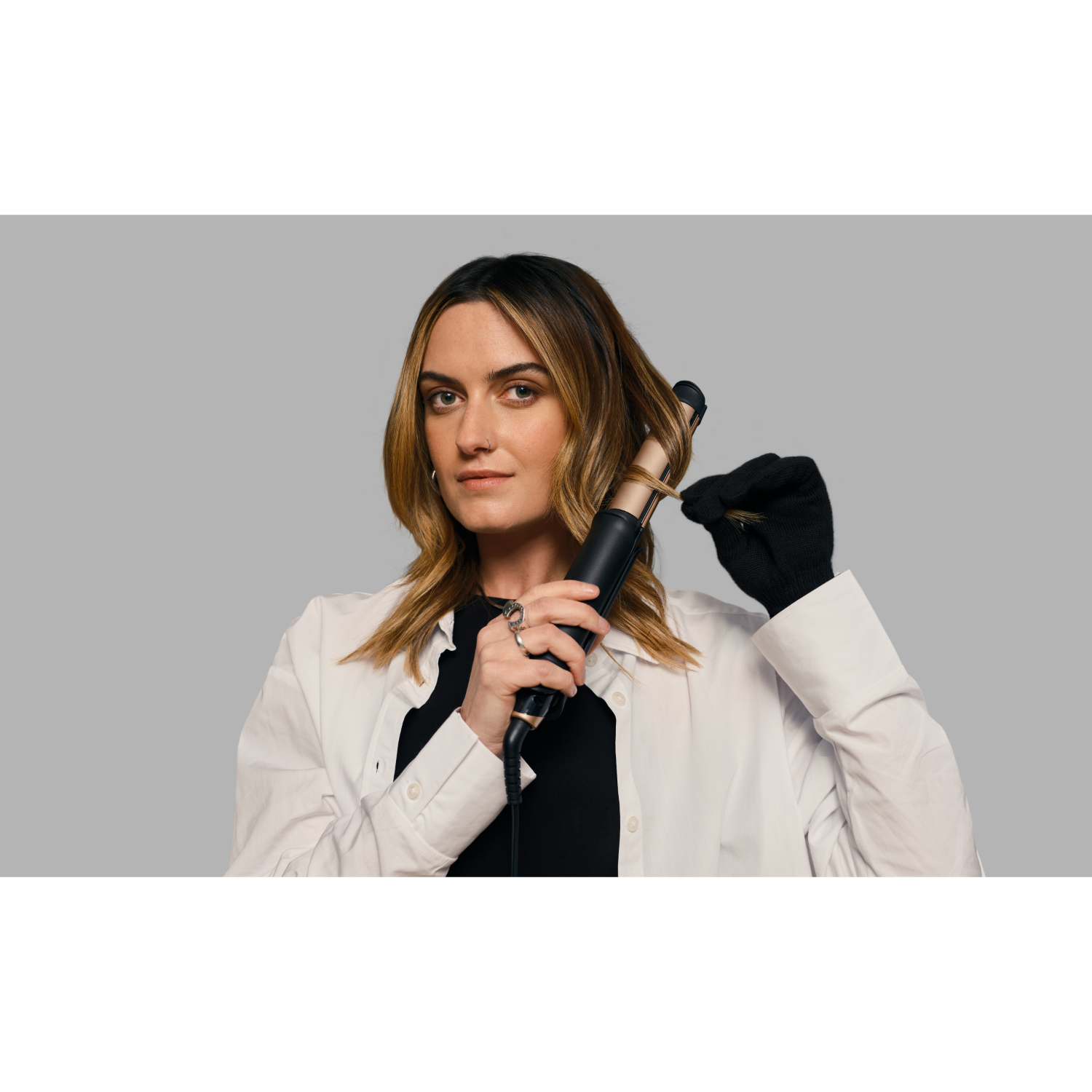 Woman with brown hair using a curling iron, wearing a white shirt, black glove and top. Gray background.