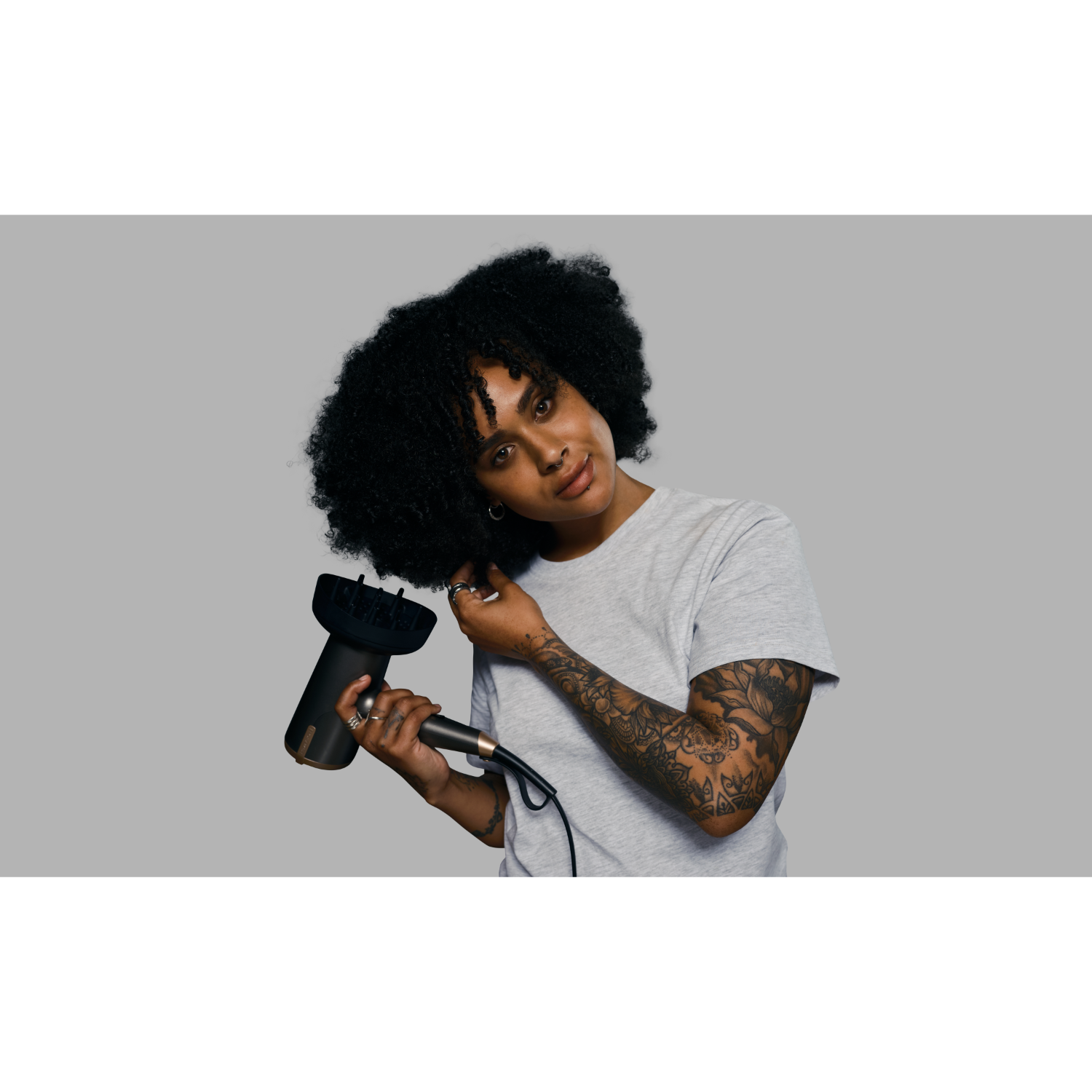 A woman with dark curly hair is holding a black hair dryer, looking towards the camera.